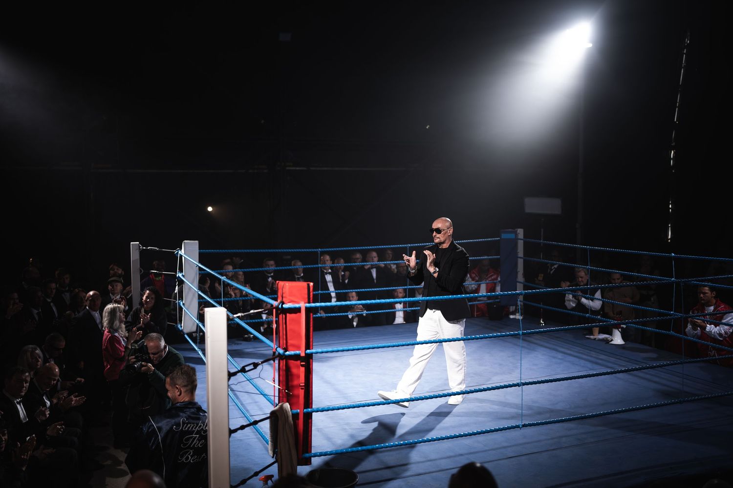 Man in white pants performs in a boxing ring under dramatic spotlight during a live event.