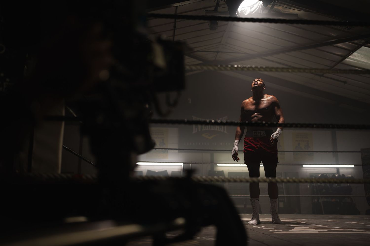 A boxer stands alone in a dimly lit boxing ring, filmed by a camera in the foreground.