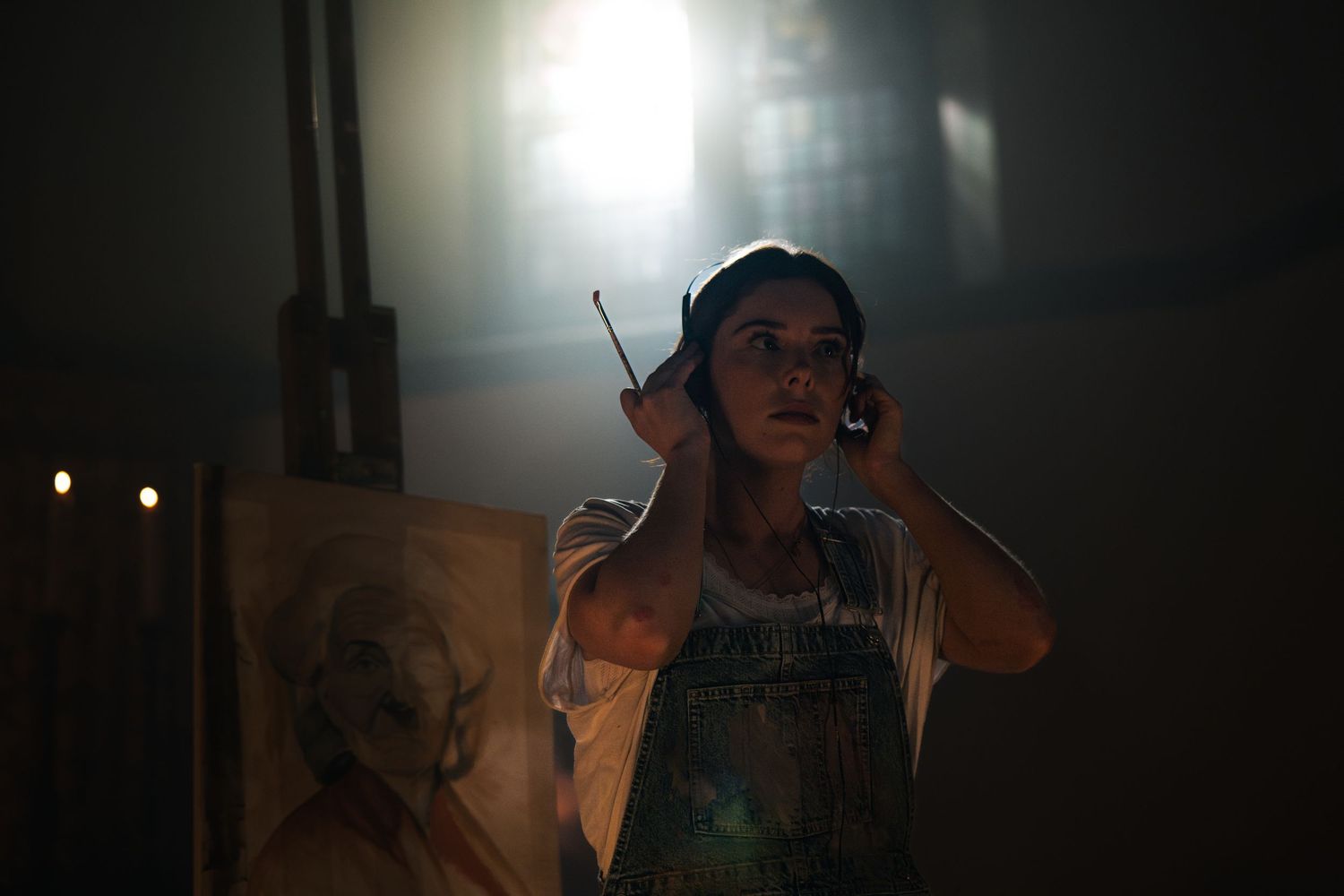 A young woman wearing headphones and overalls stands in a dimly lit studio near a canvas painting.