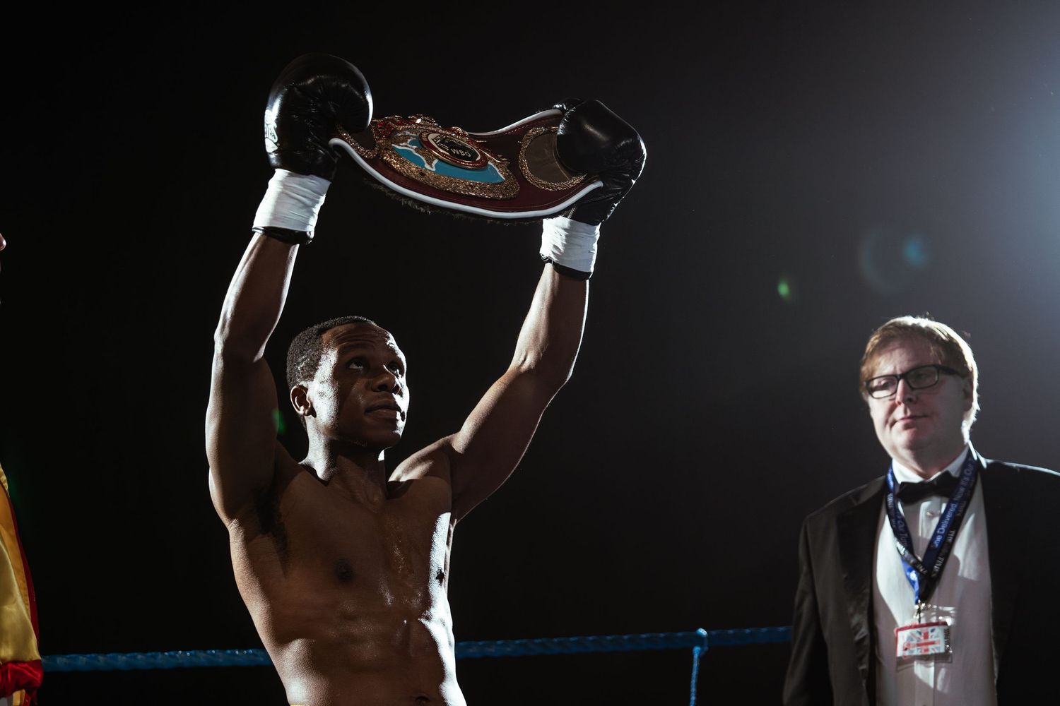 Triumphant boxer raises championship belt overhead in ring, wearing black gloves, while official in tuxedo stands nearby.
