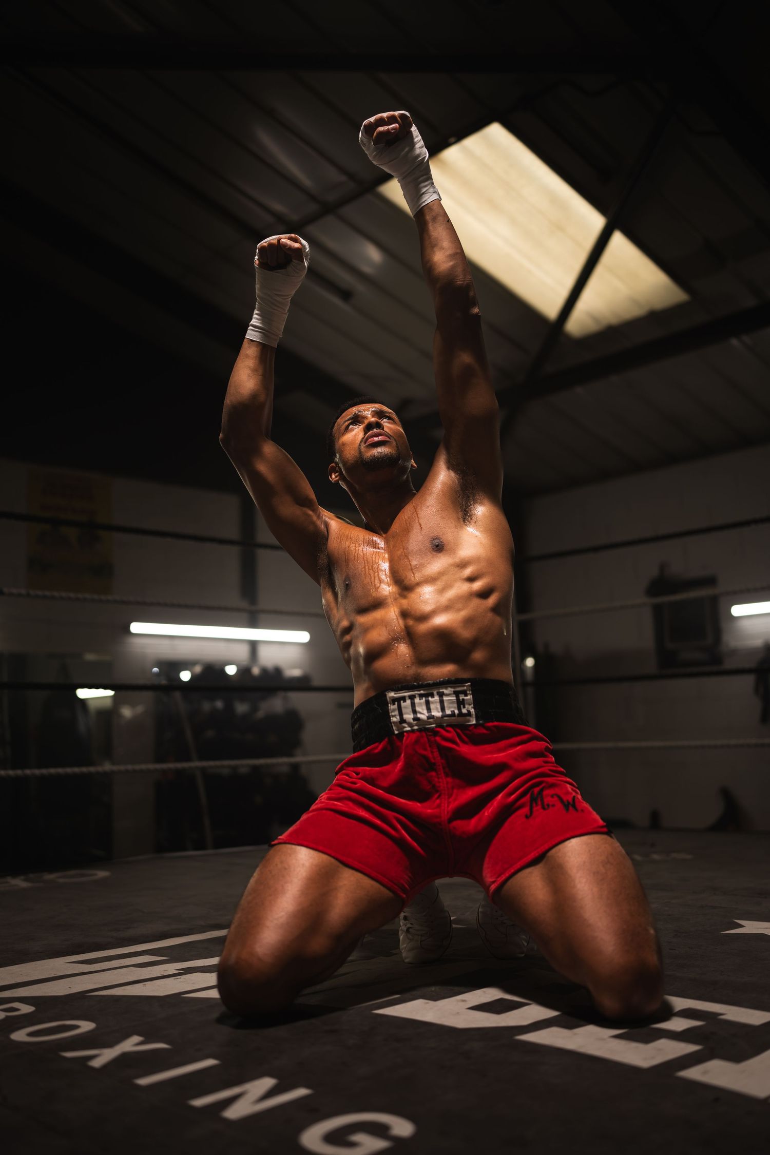 A muscular shirtless boxer kneels in a boxing ring with arms raised in victory.