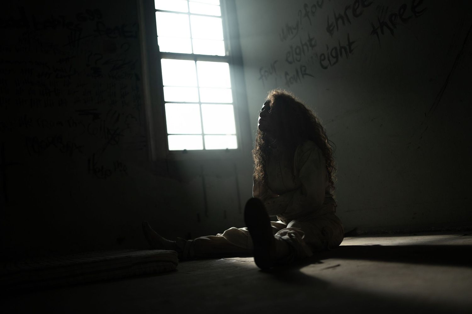Silhouette of a person sitting alone in a dark room with writings on the wall, illuminated by a single window.