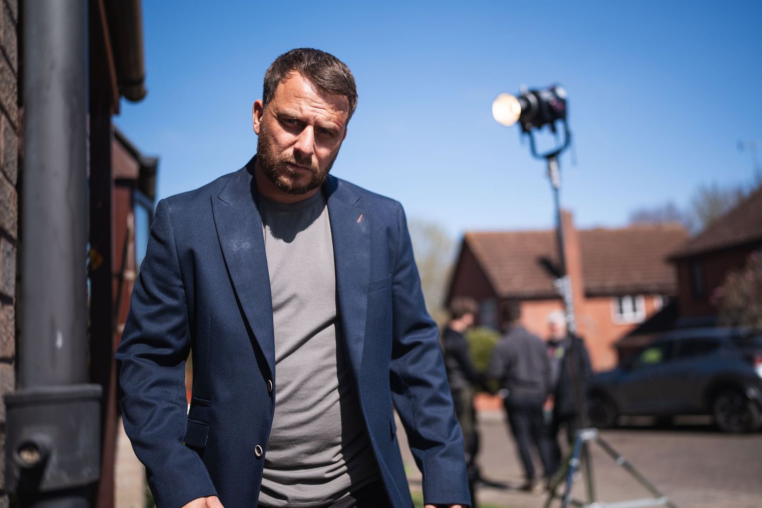 A man in a navy blazer walks outdoors on a film set on a sunny day.