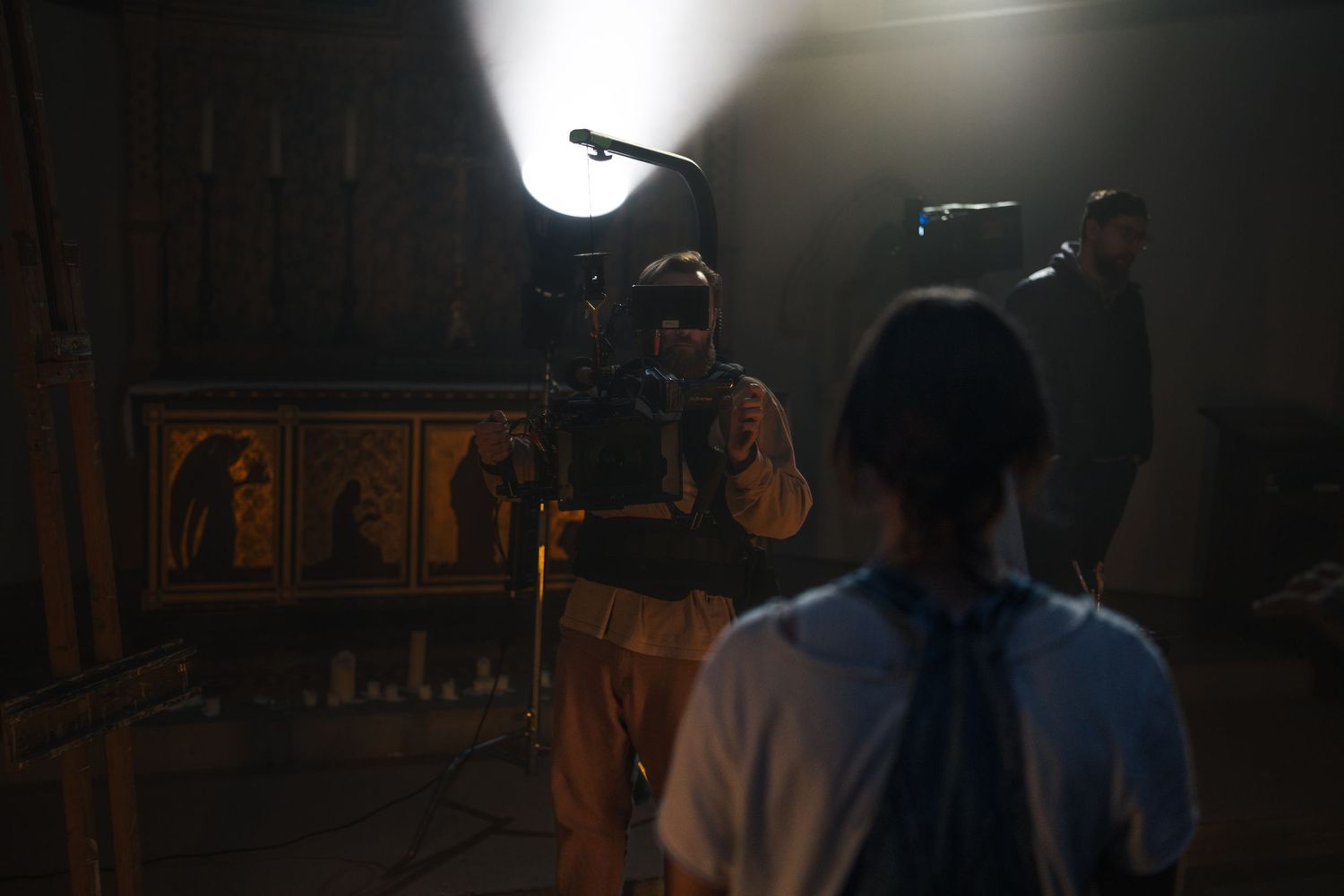 Behind-the-scenes film set with a camera operator filming a scene lit by a large studio light in a dark room.