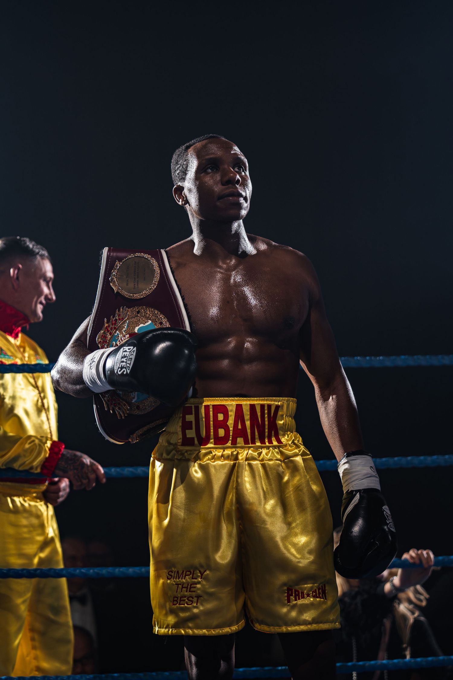 A boxer in gold Eubank shorts holds a championship belt inside a boxing ring.