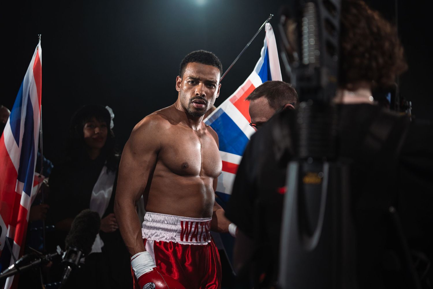 A shirtless boxer in red shorts stands before a Union Jack flag at a boxing event.