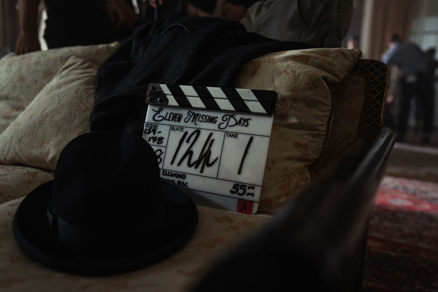 A film clapperboard rests on a couch beside a black fedora hat on a dimly lit movie set.