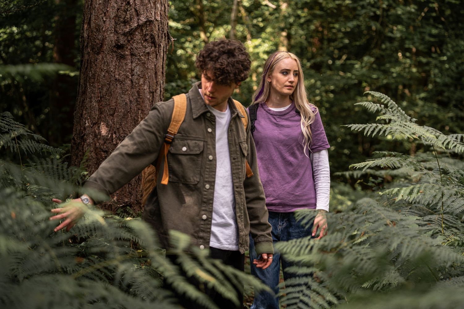 Young man and woman explore a lush green forest with ferns and towering trees around them.