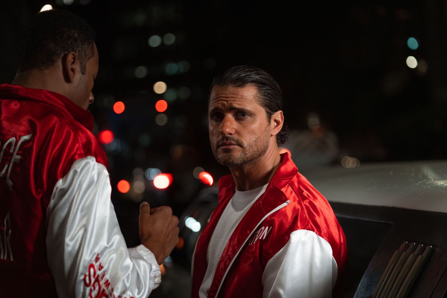 Two men in matching red and white track jackets having a serious conversation outdoors at night.