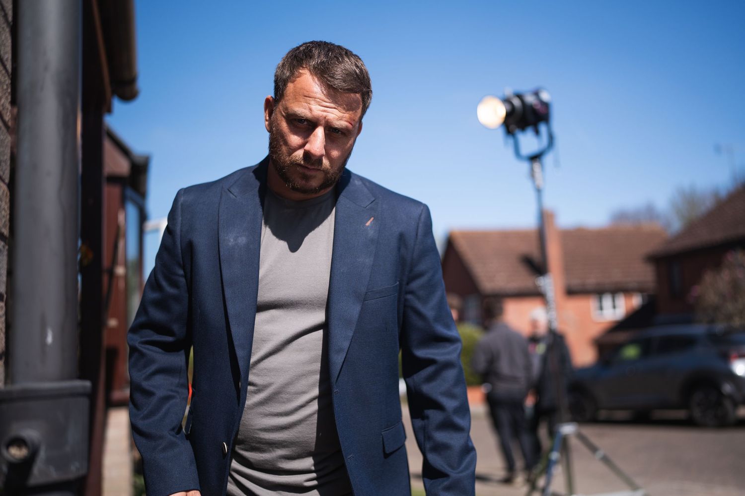 A man in a blue blazer and grey shirt walks outdoors on a sunny day with film equipment visible behind him.