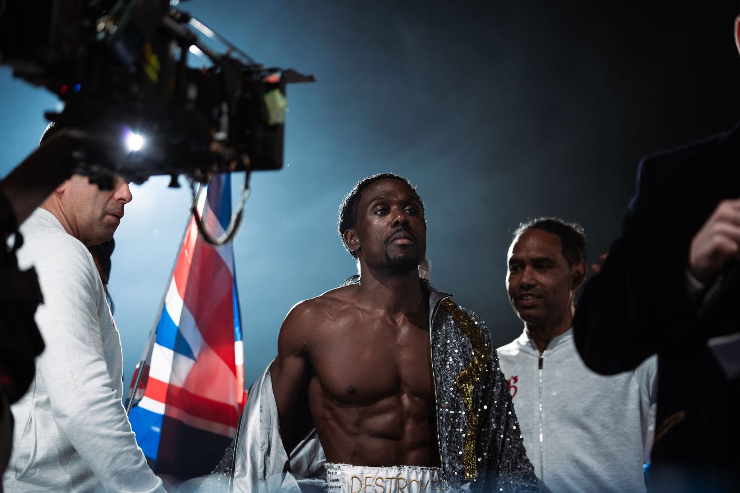 Shirtless boxer entering arena with British flag cape and support team.