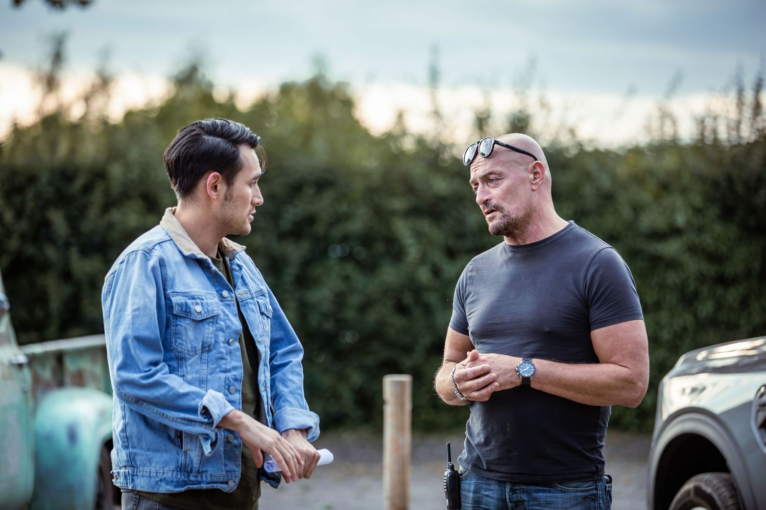 Two men having an outdoor conversation, one in a denim jacket and one in a grey t-shirt, standing near green hedges at dusk.