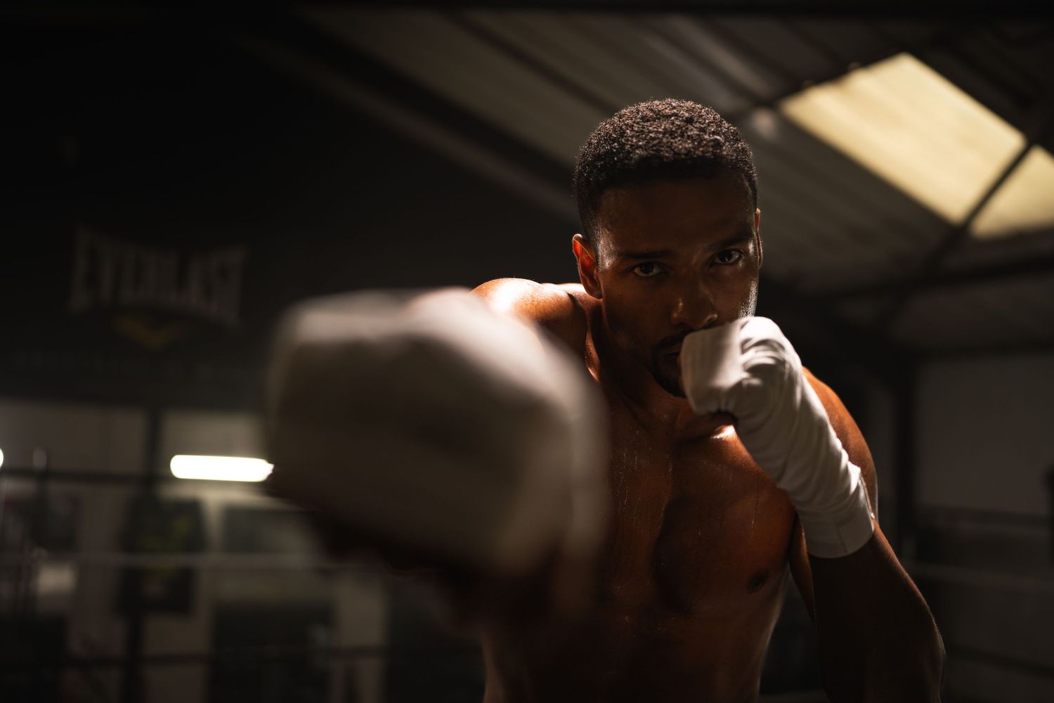 A focused young boxer in a gym throws a punch toward the camera while wearing white hand wraps.