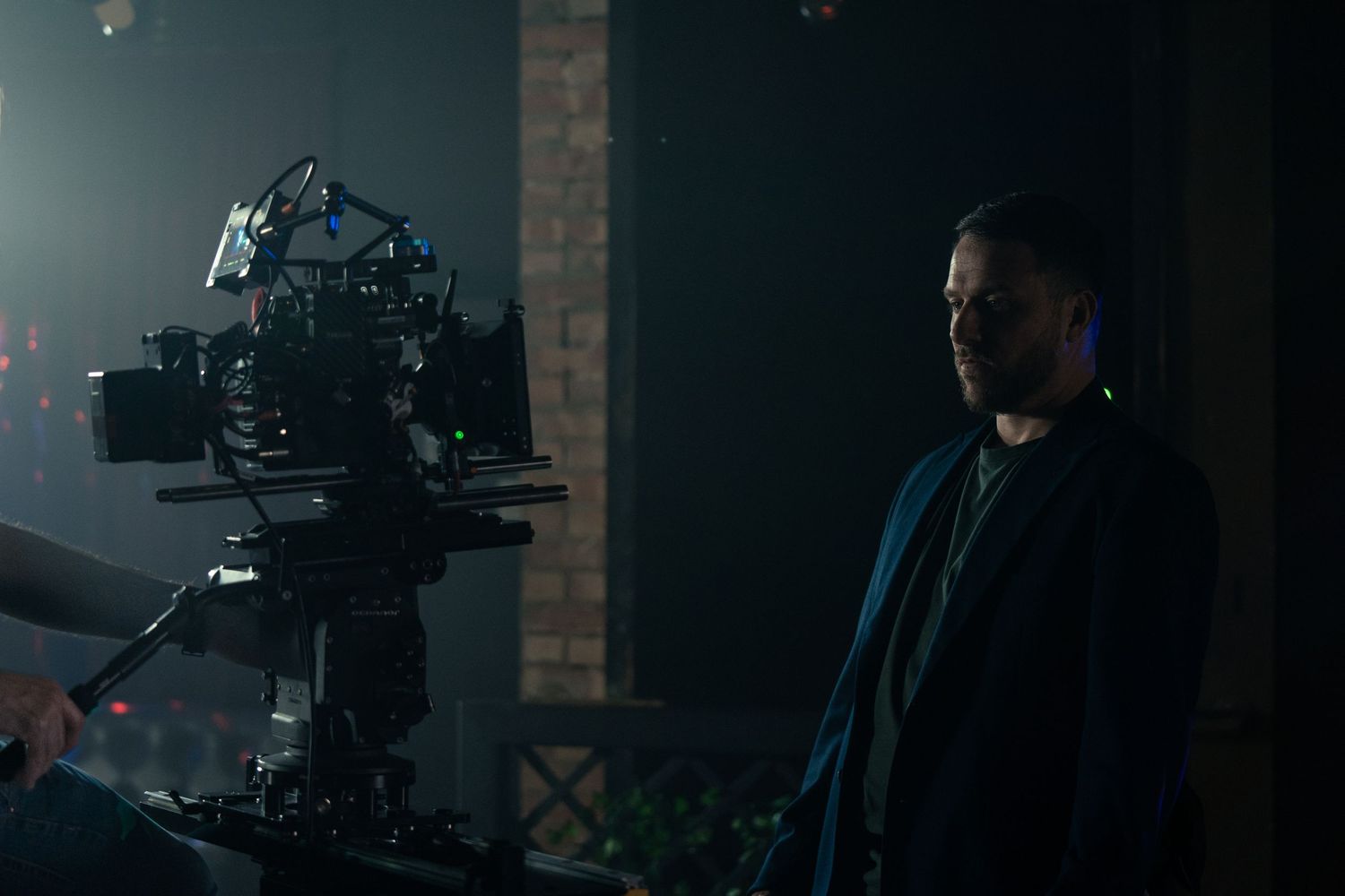 A bearded man in a dark jacket stands beside a professional film camera during a nighttime production shoot.
