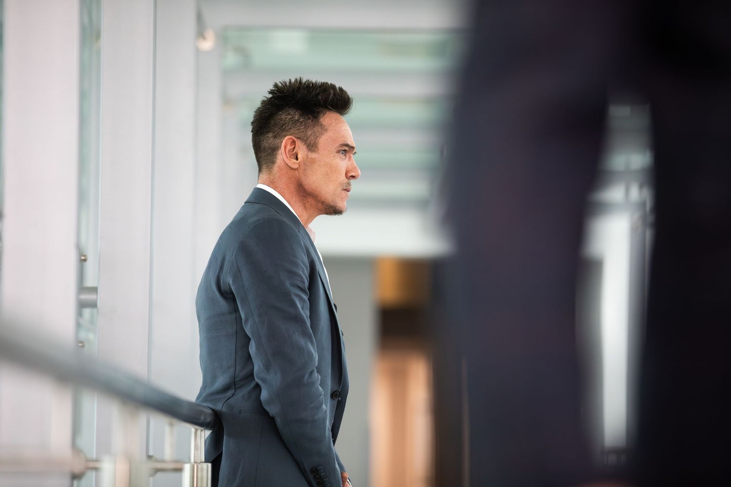 A man in a grey suit stands in profile in a modern building interior.
