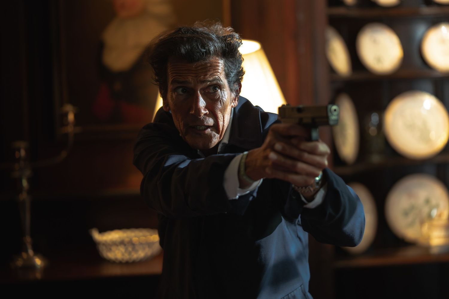 An older man aims a handgun in a dimly lit, elegantly furnished room.