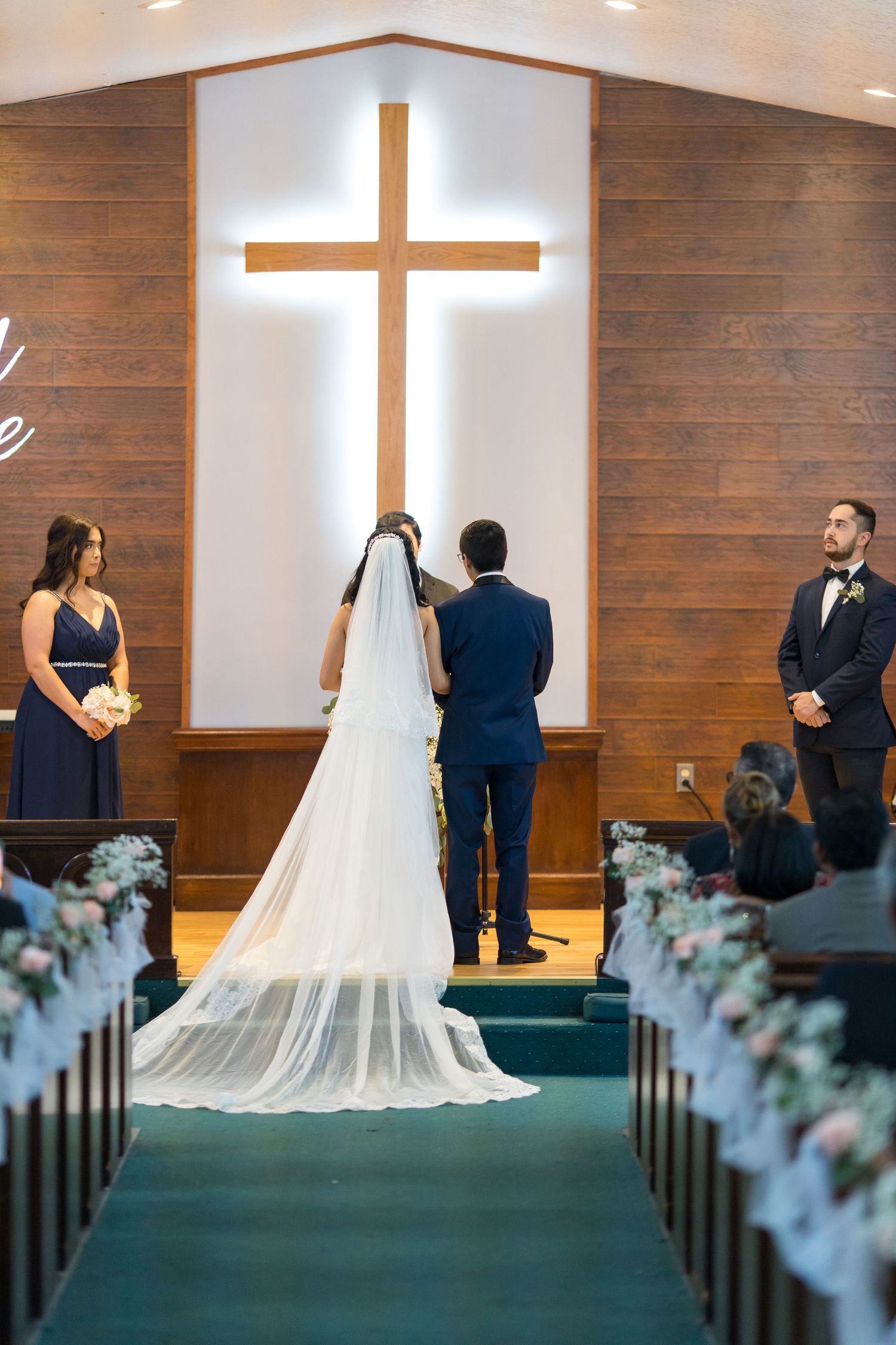 Bride and groom stand at church altar before illuminated cross, flanked by bridesmaid and groomsman during wedding ceremony.
