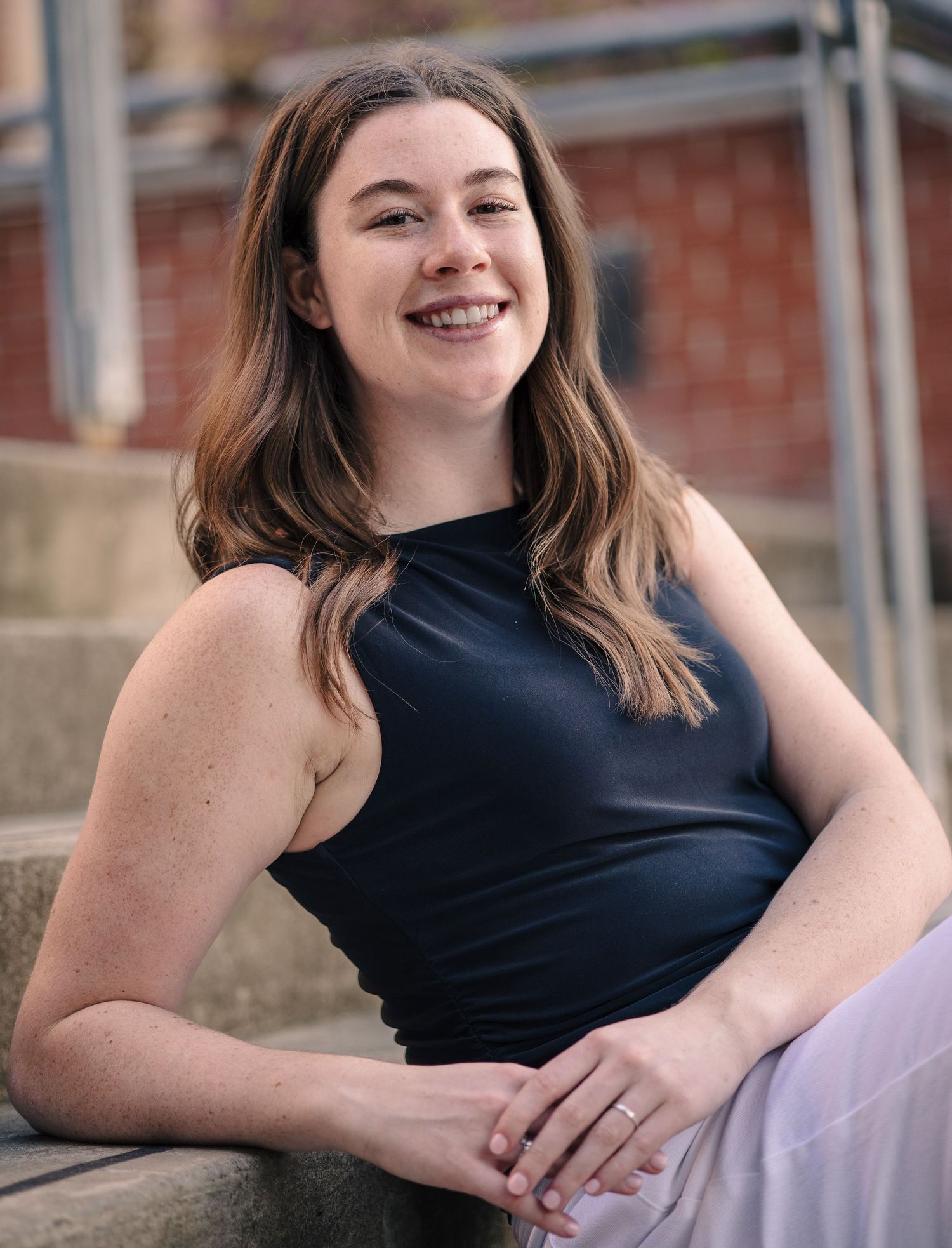 Smiling young woman with dark hair and freckles sitting on steps wearing a black sleeveless top.