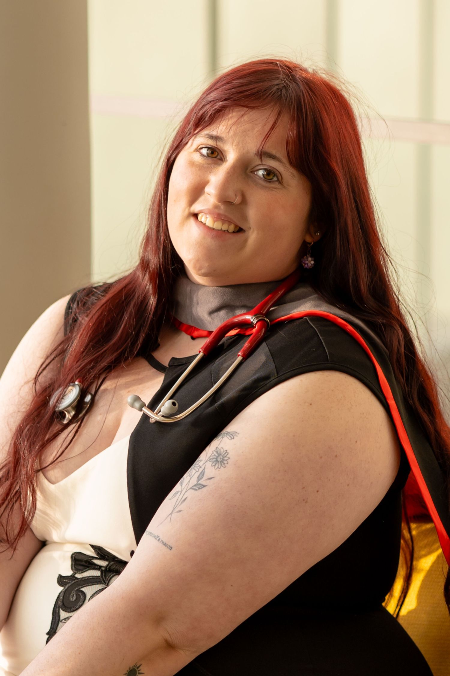 Smiling woman with red hair wearing a stethoscope and graduation gown, showing tattoos on her arm.