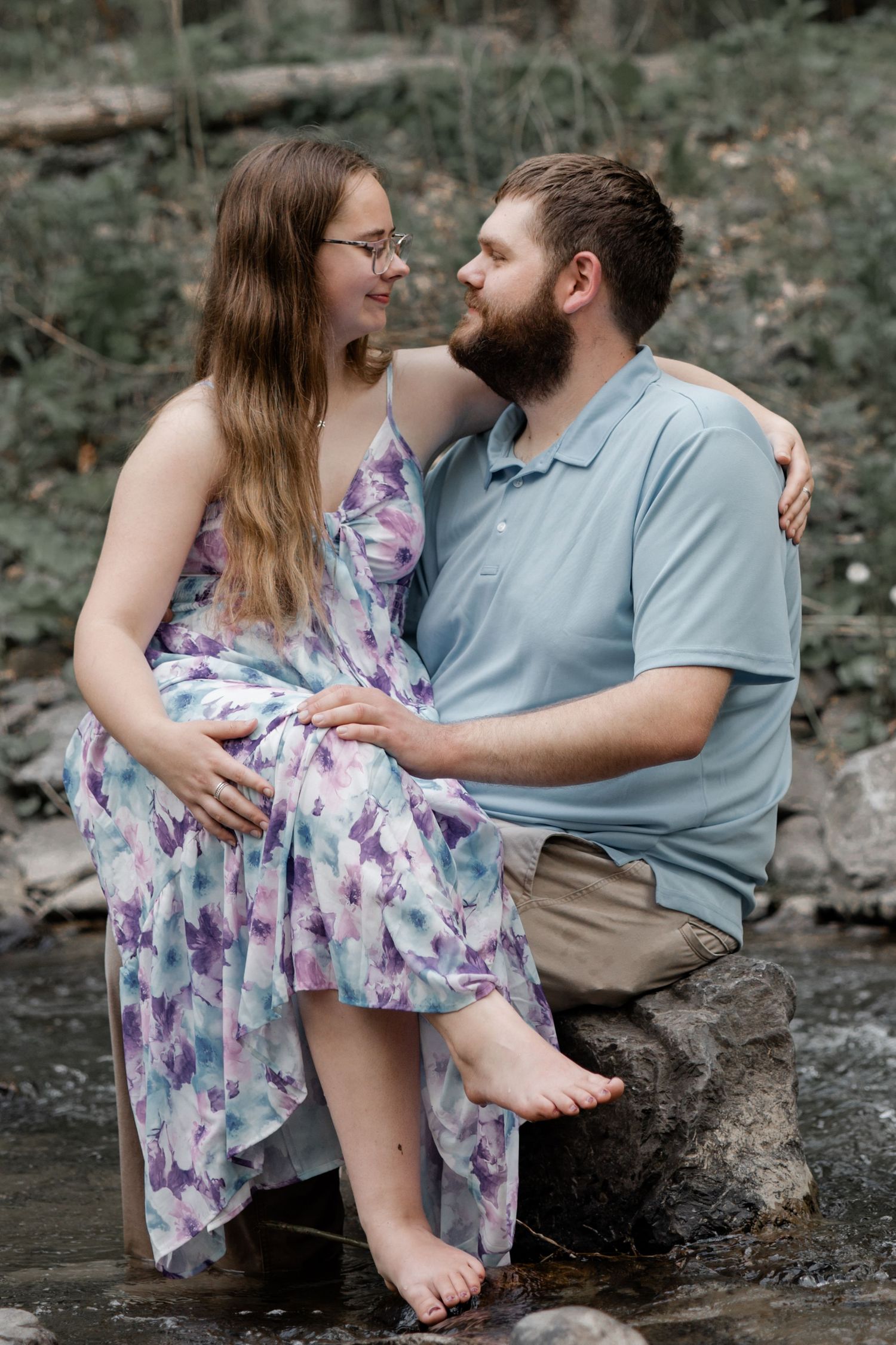 Couple sitting on creek rocks, woman in purple floral dress and man in blue polo smiling at each other.