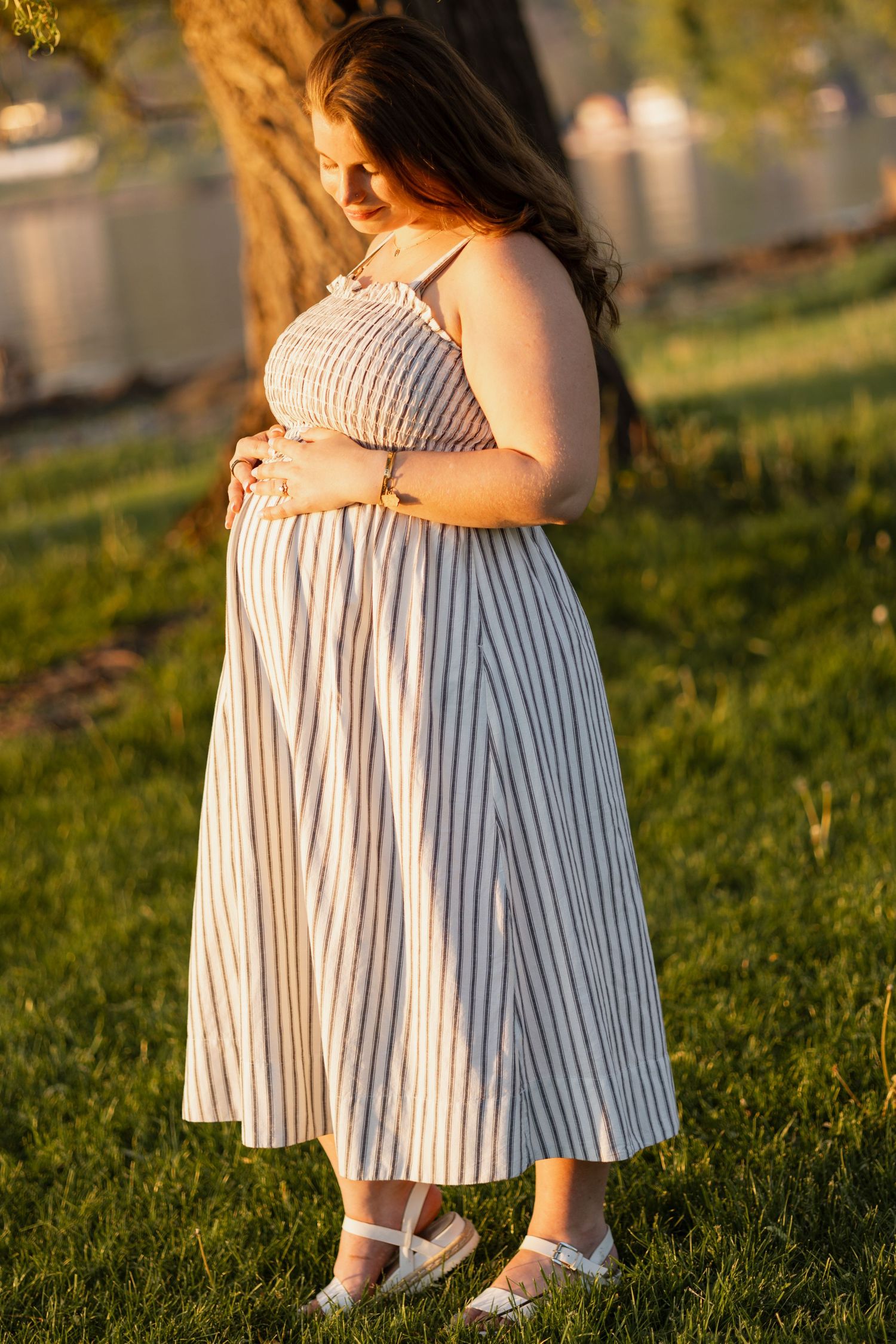 Pregnant woman in striped sundress cradling baby bump in golden hour sunlight.