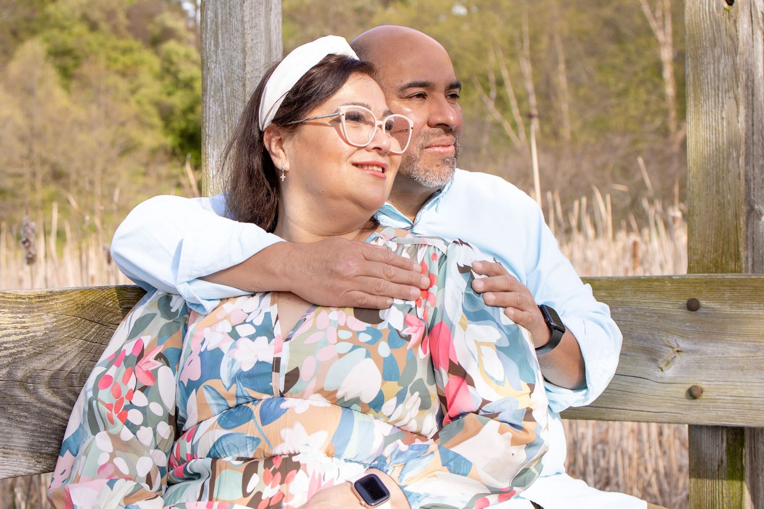 Couple sitting on a wooden bench outdoors, woman in floral top with glasses, man in light blue shirt smiling.