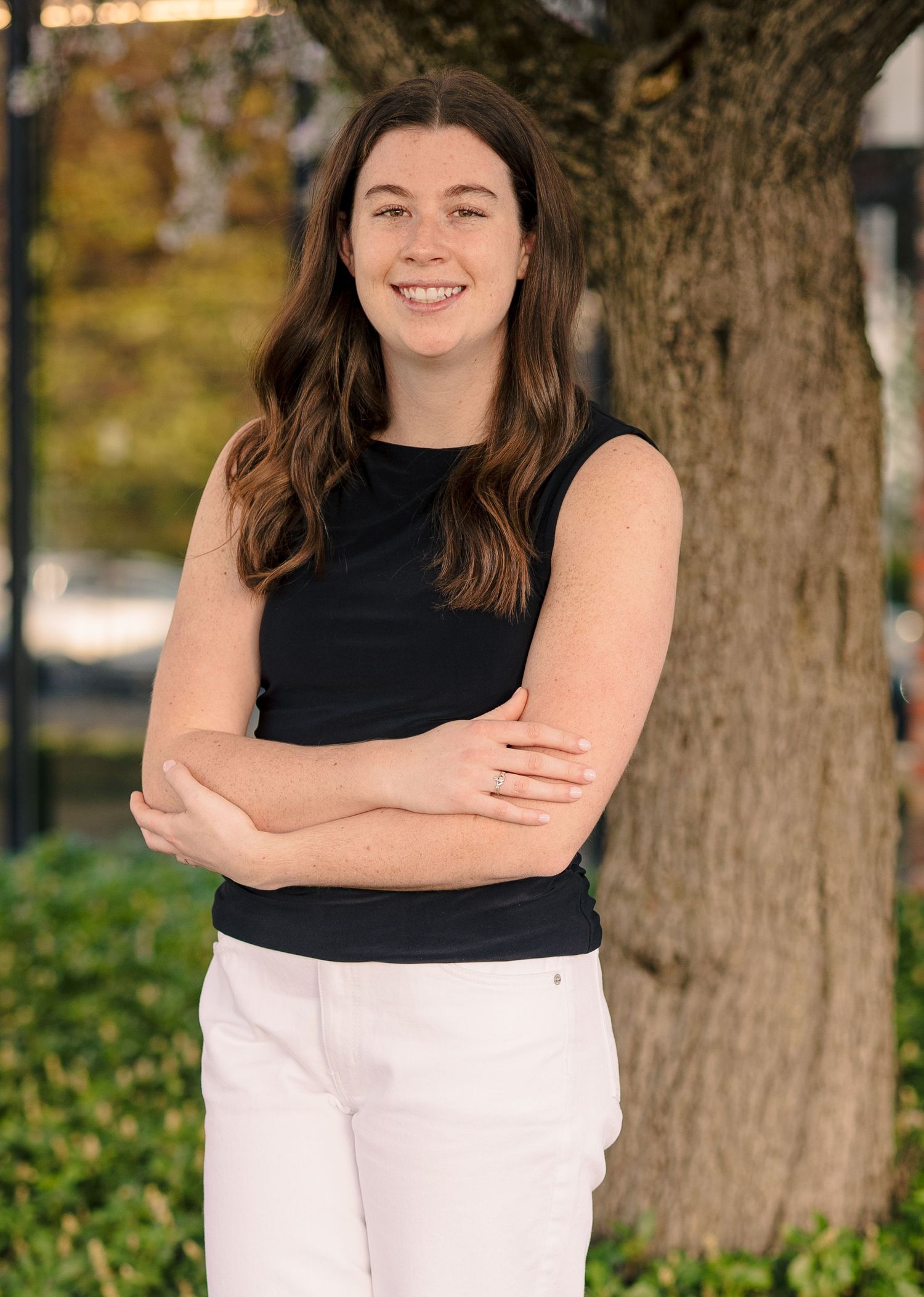 Young woman with dark wavy hair in black sleeveless top and white pants standing by a tree outdoors.