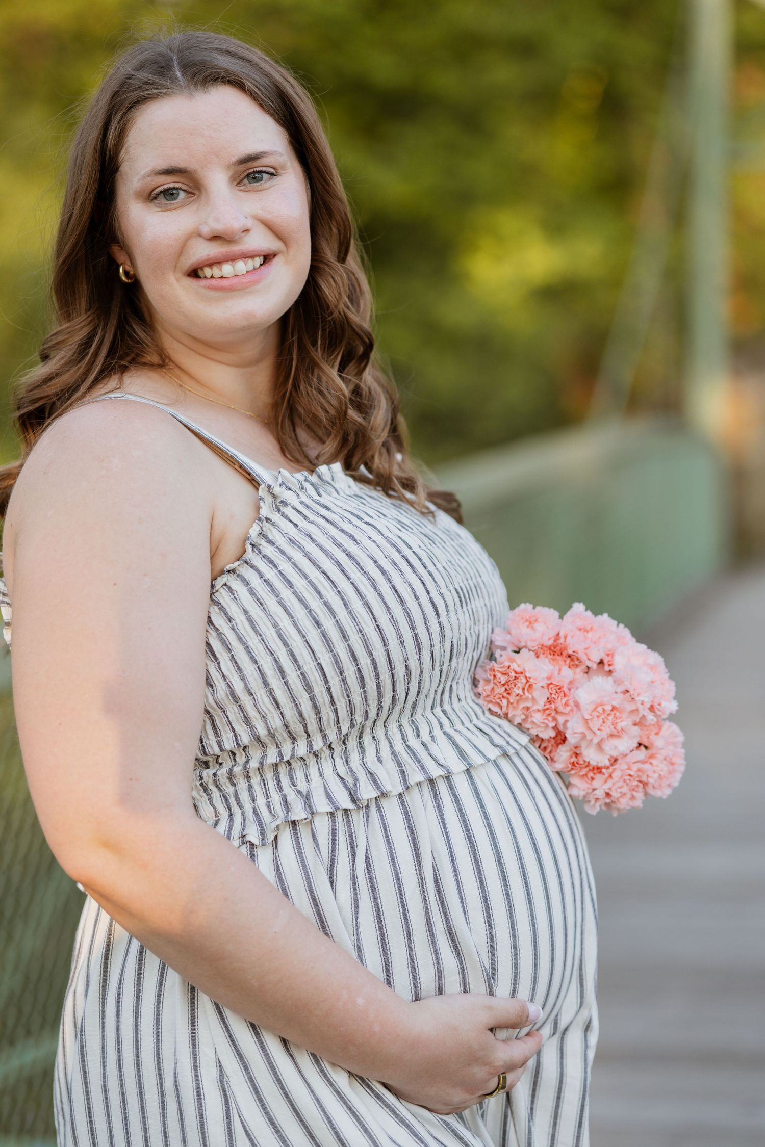 Smiling pregnant woman in striped sundress holding pink carnations outdoors on a bridge surrounded by green trees.