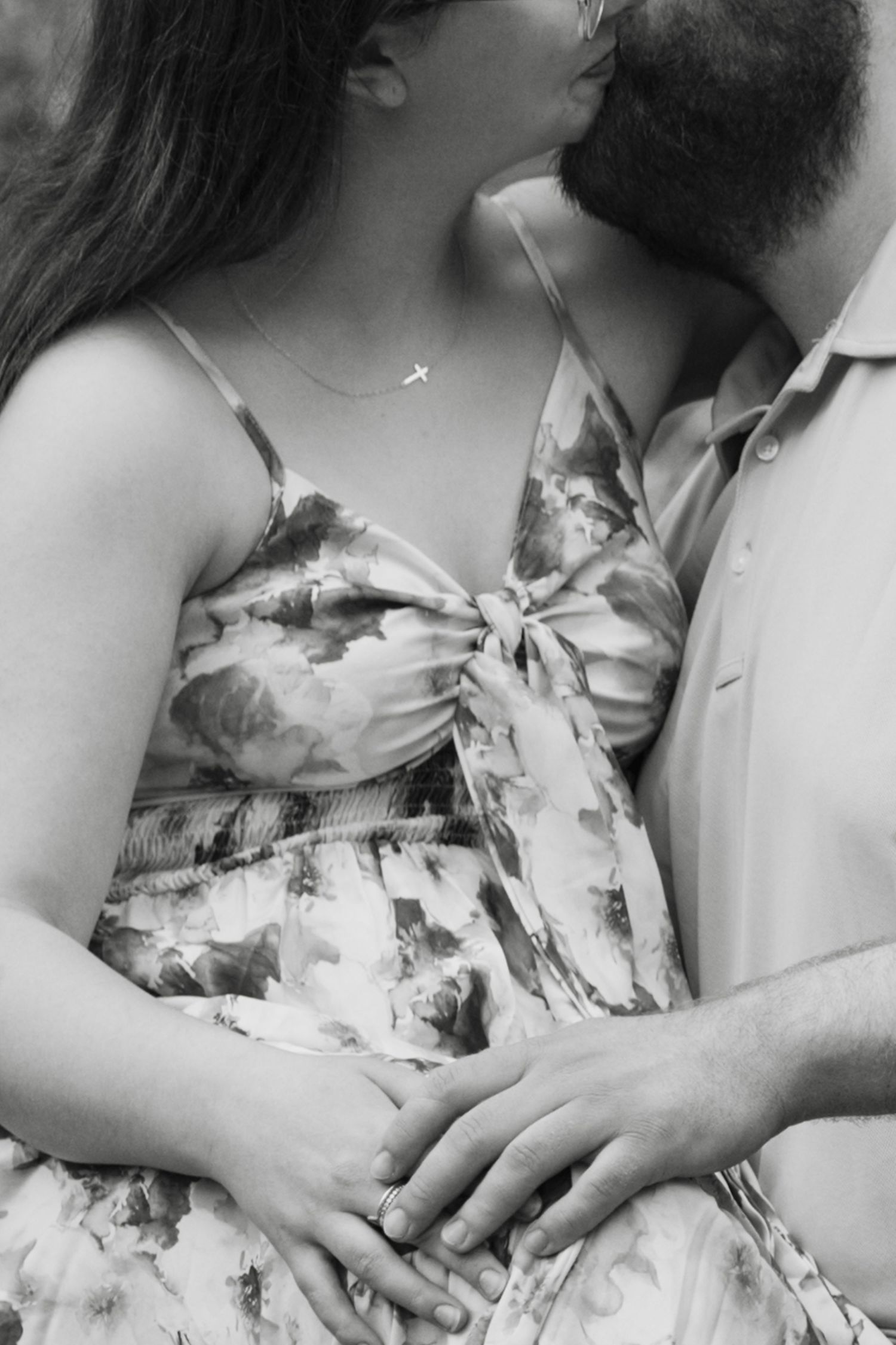 Black and white photo of a couple kissing, woman in floral dress with cross necklace, hands intertwined at her waist.