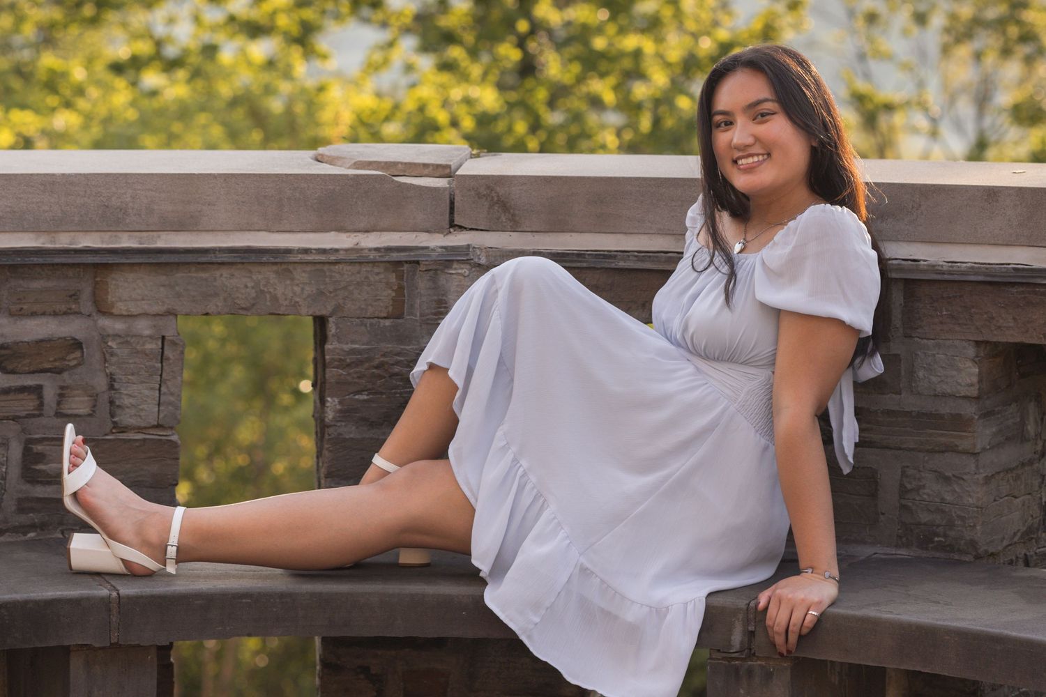 Young woman in white dress sitting on stone bridge railing, smiling outdoors with green trees in background.