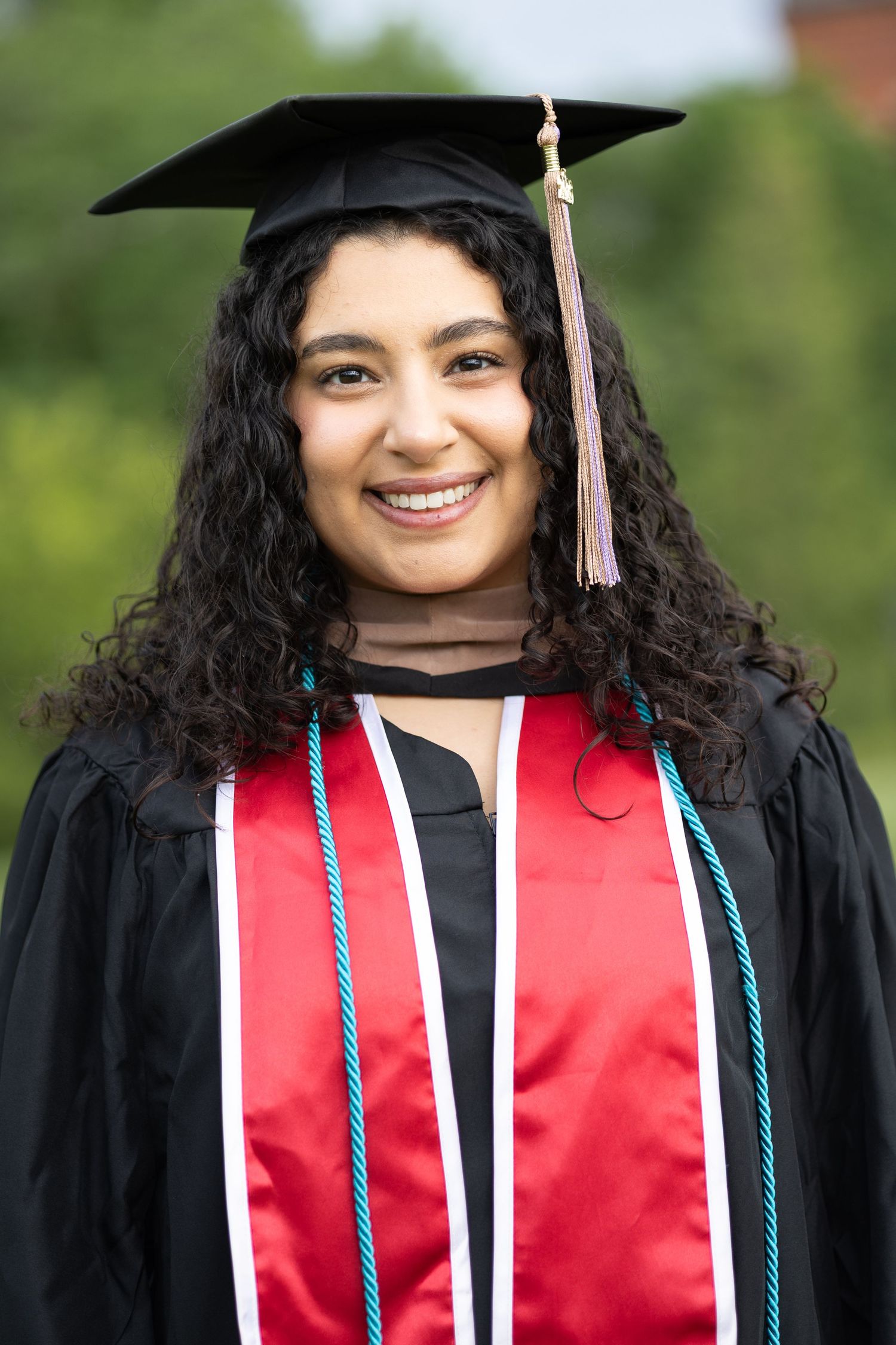 Smiling young woman in black graduation cap and gown with red stole and blue cord outdoors.