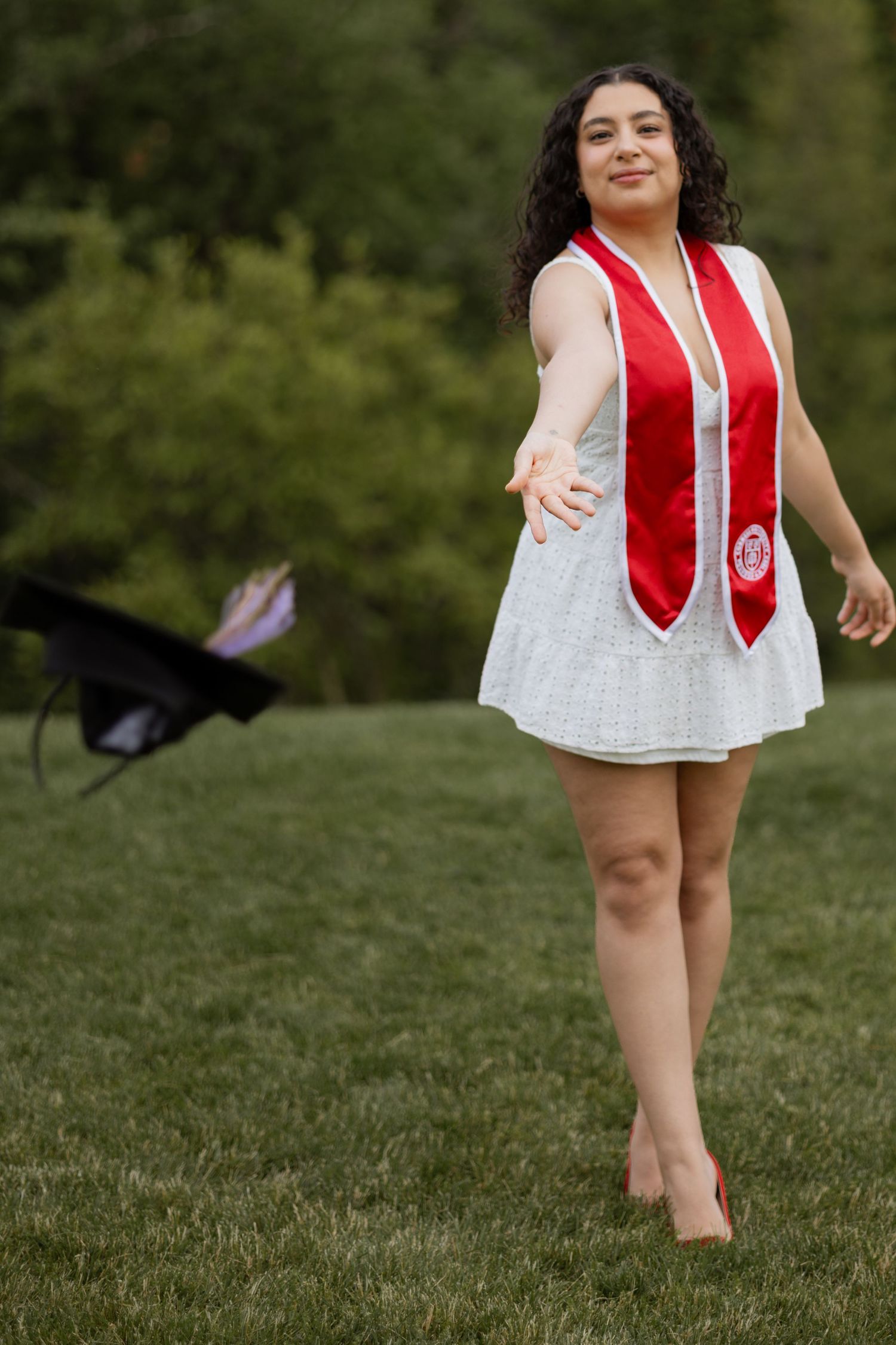 A young woman in a white dress and red graduation stole tosses her cap on a green lawn.