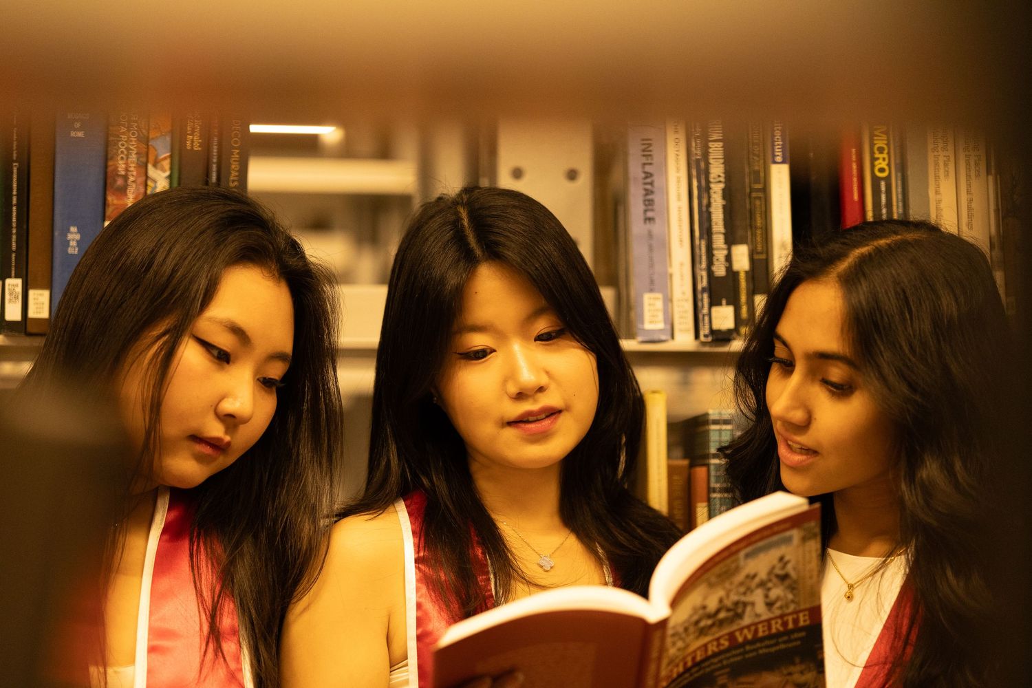 Three young women reading a book together in a library with bookshelves in the background.