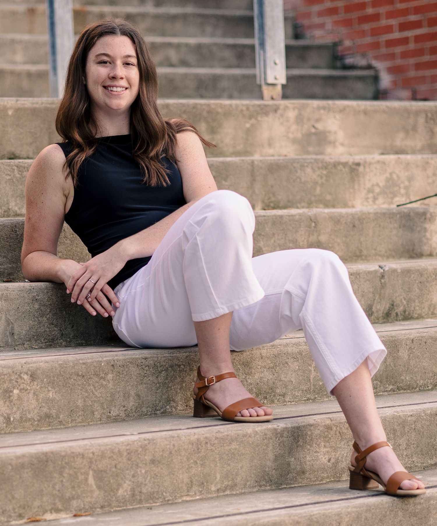 Young woman in white pants and black top sitting on stone steps, smiling, wearing brown sandals.
