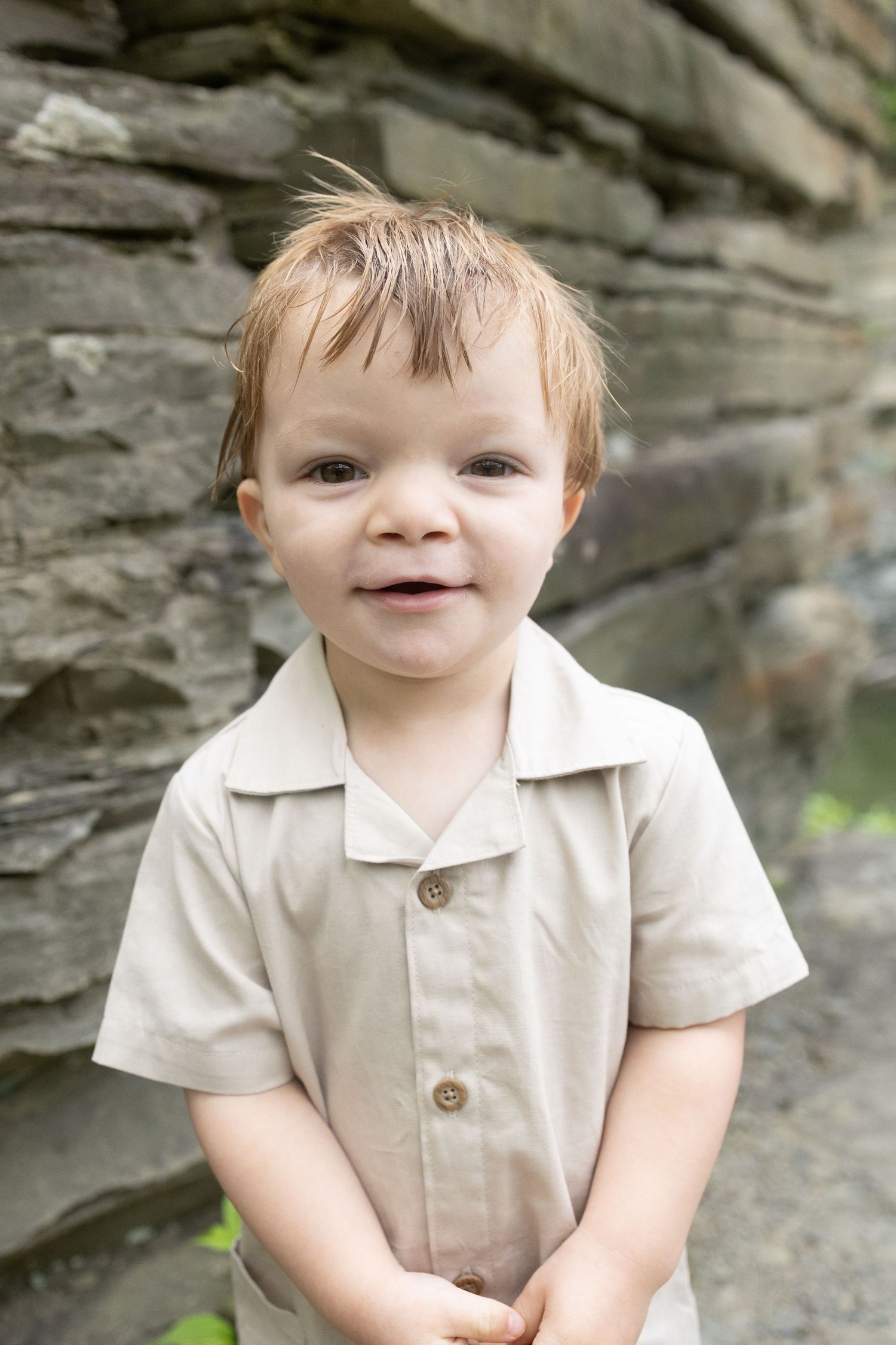 Smiling toddler boy with light brown hair wearing a beige button-up shirt standing in front of a stone wall.