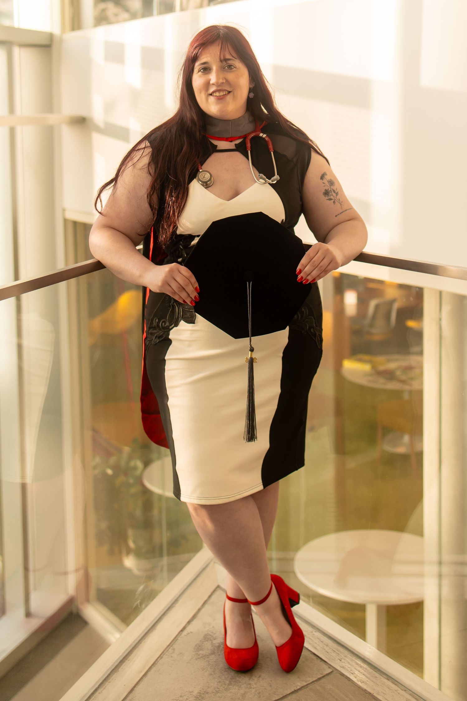 Woman in black and white dress with red heels and cape poses confidently on indoor balcony.