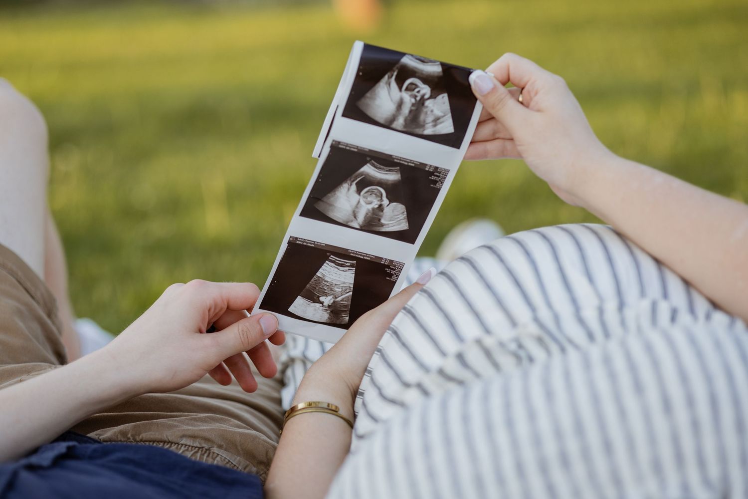 Pregnant woman holding ultrasound sonogram photos while relaxing on green grass outdoors.