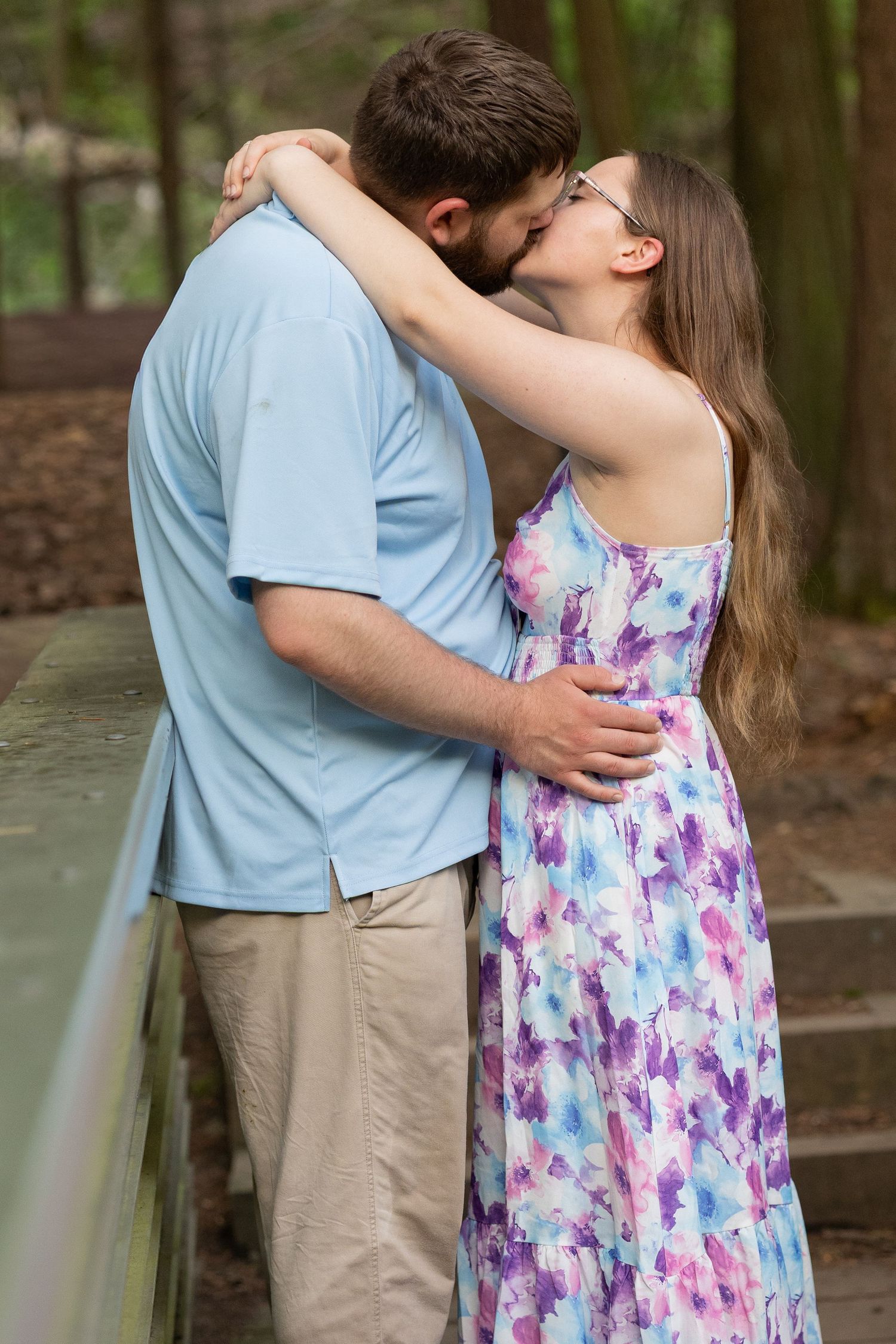A couple shares a tender kiss outdoors, woman in floral dress embracing bearded man in blue shirt near wooden railing.