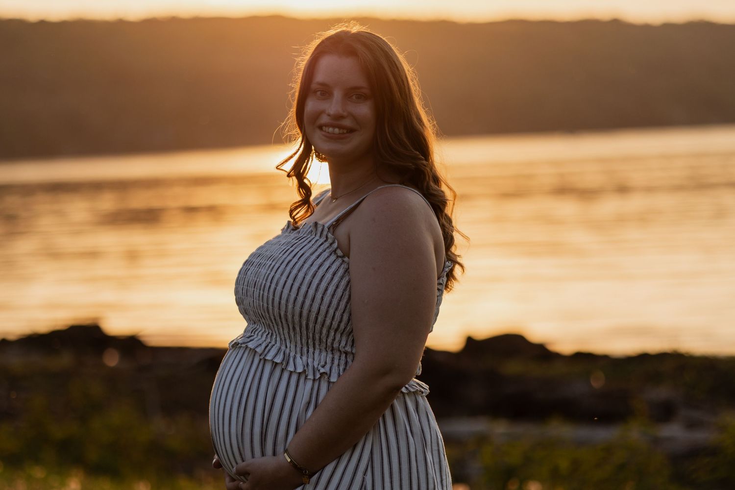Pregnant woman in striped dress smiling at sunset by the water, golden hour maternity portrait.