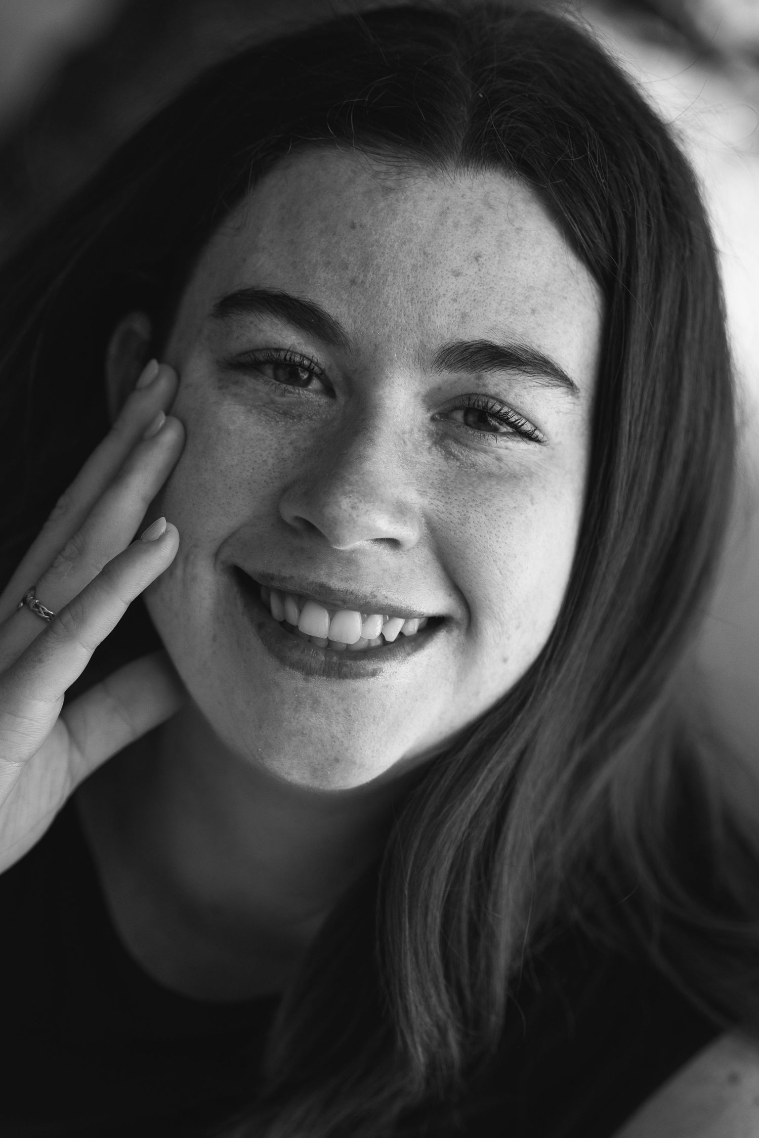 Black and white portrait of young smiling woman with freckles and dark hair touching her cheek.