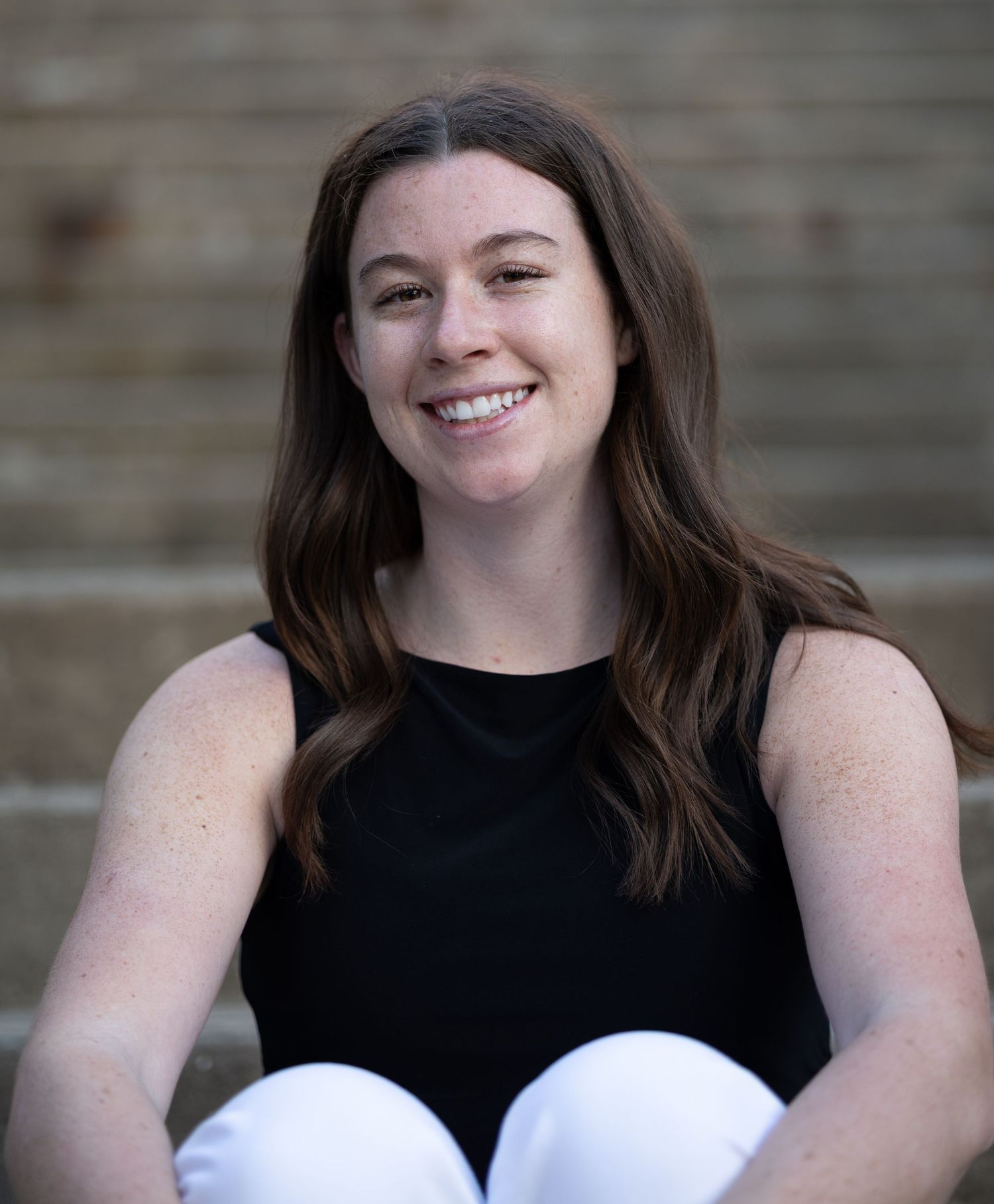 Smiling young woman with brown hair wearing a black sleeveless top seated outdoors on steps.