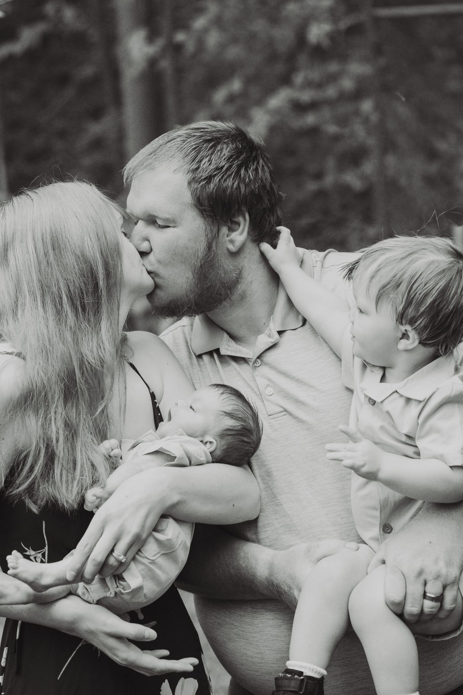 Black and white photo of parents kissing while holding newborn baby and toddler outdoors in nature.