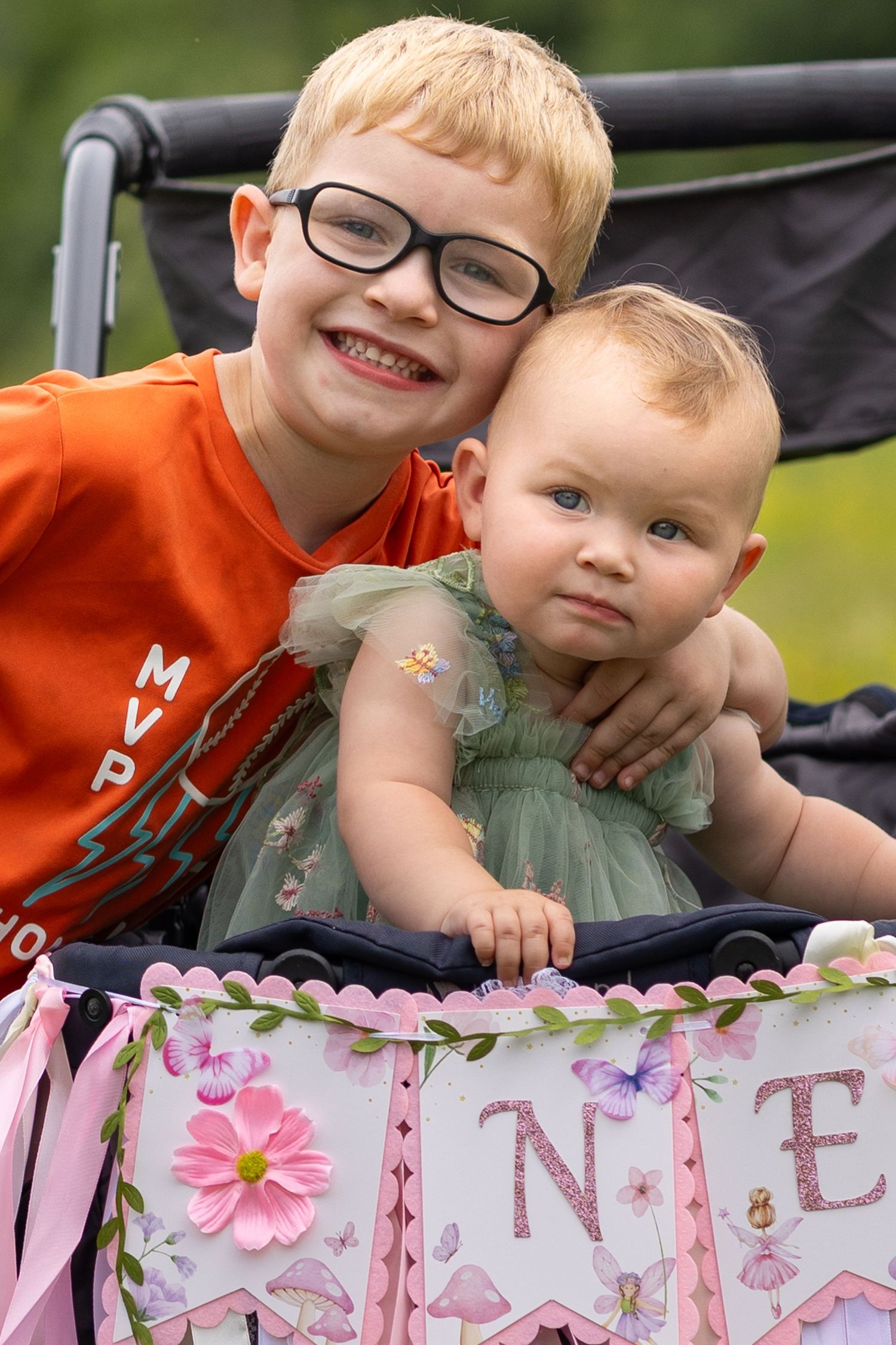 A smiling boy with glasses in an orange shirt hugs a baby girl in a green dress at a first birthday party.