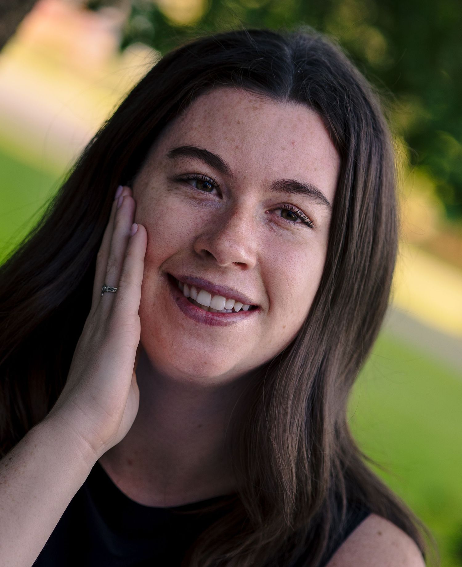 Young woman with freckles and long brown hair smiling warmly outdoors with green bokeh background.