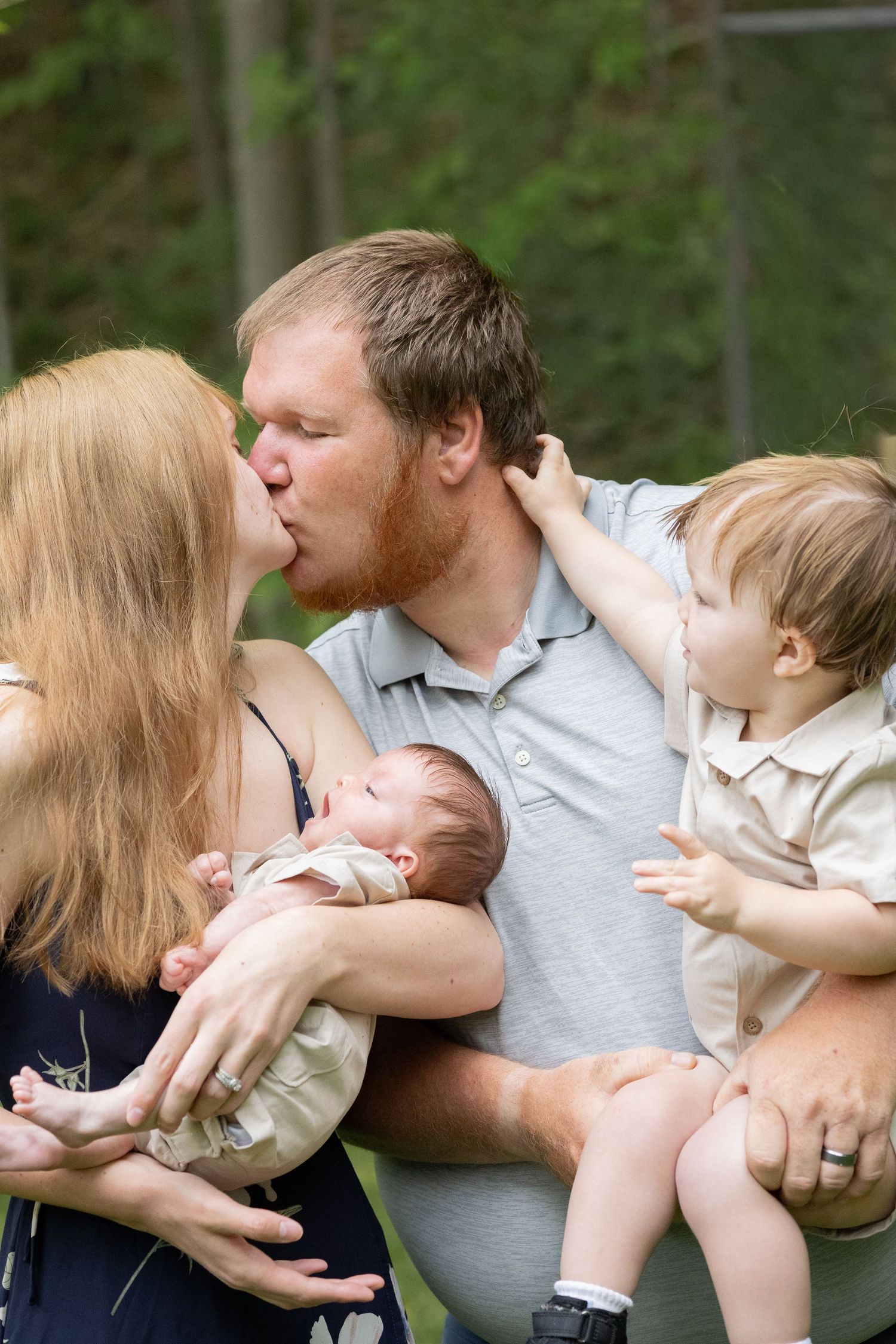 A couple kisses while holding a newborn and toddler outdoors, capturing a sweet family moment.