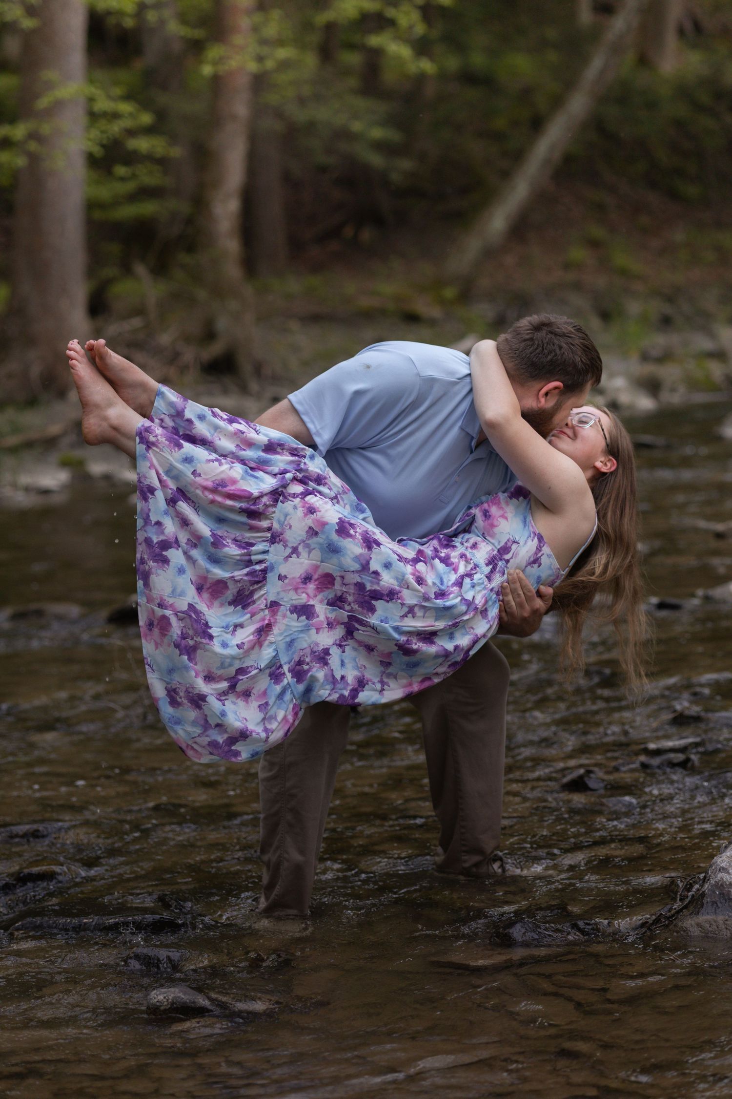 A romantic couple shares a kiss in a forest creek, man lifting woman in a floral dress, surrounded by lush green trees.