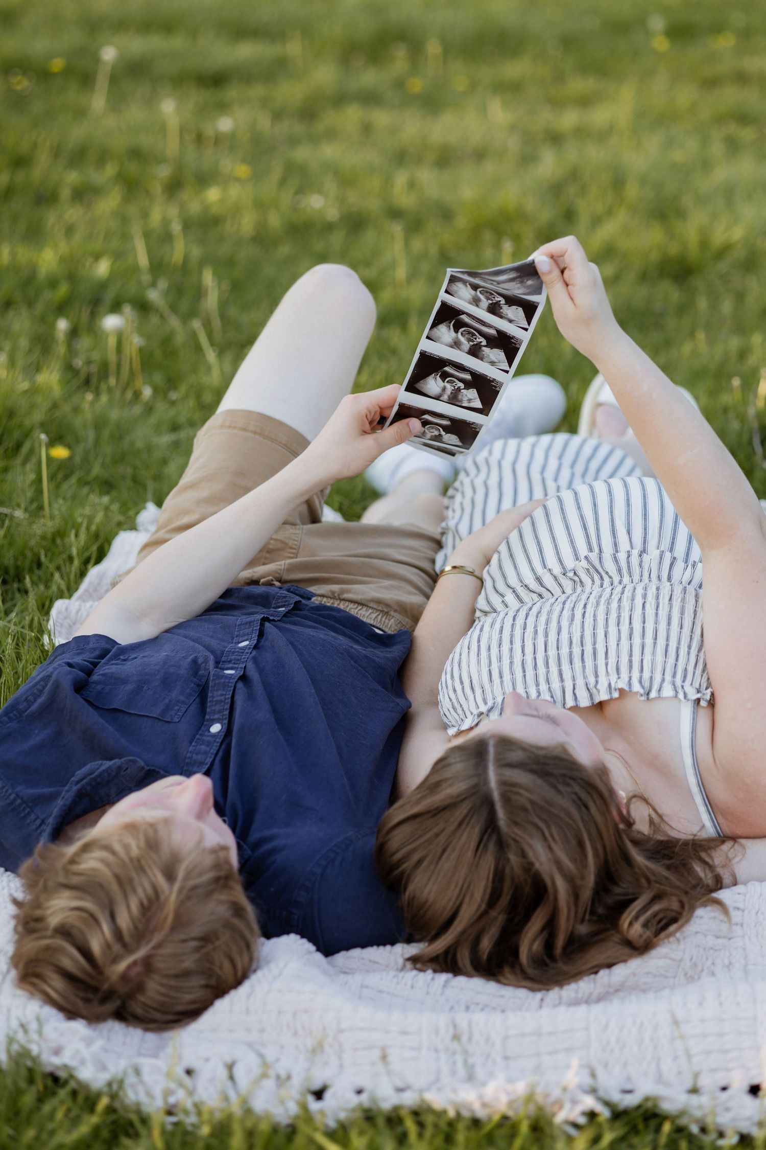 Expecting couple lying on grass holding ultrasound photos above them on a white blanket.