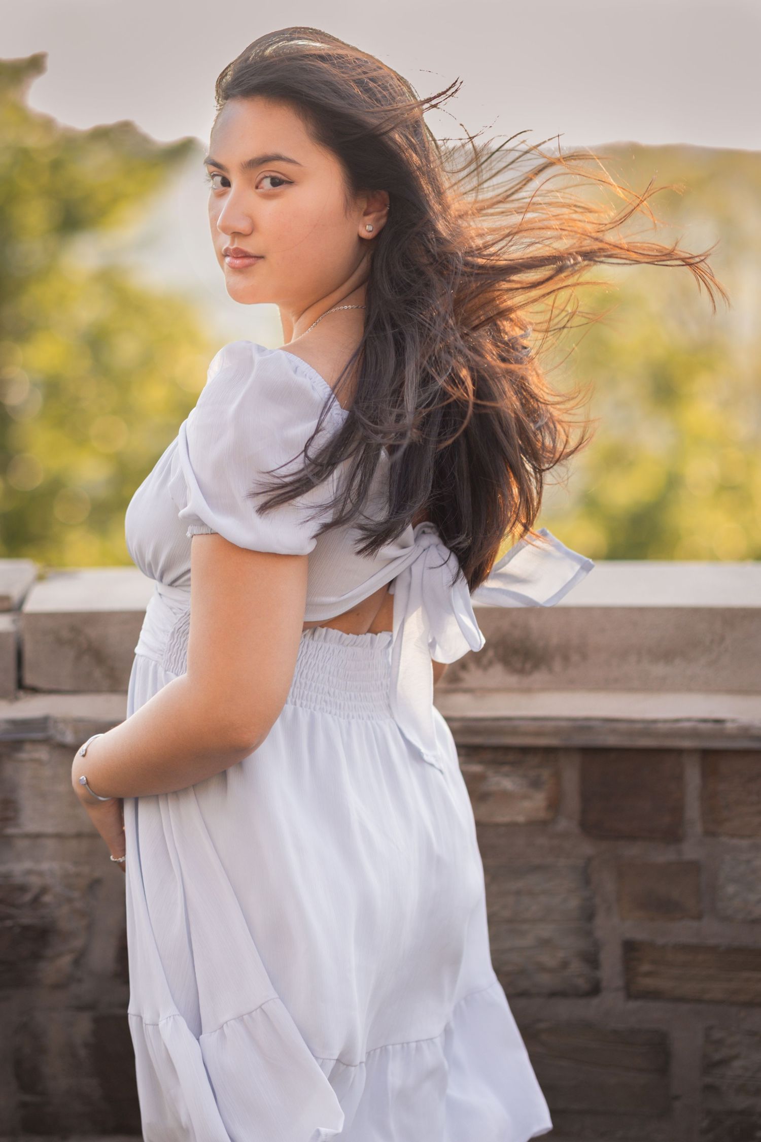 Young woman in white ruffled dress with wind-blown dark hair poses outdoors near stone wall at sunset.