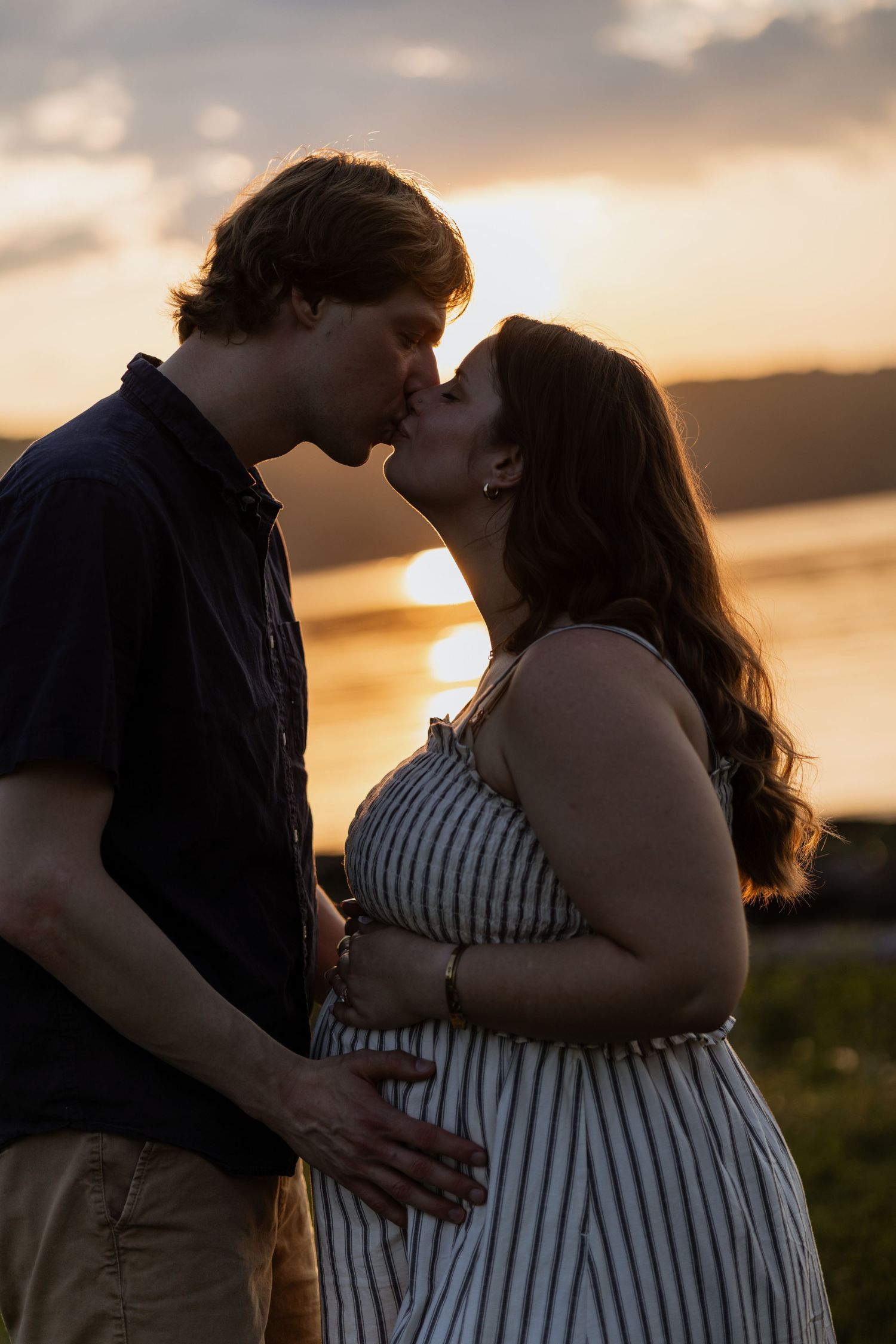 Couple sharing a kiss at sunset, woman in striped dress with visible baby bump near a lake.