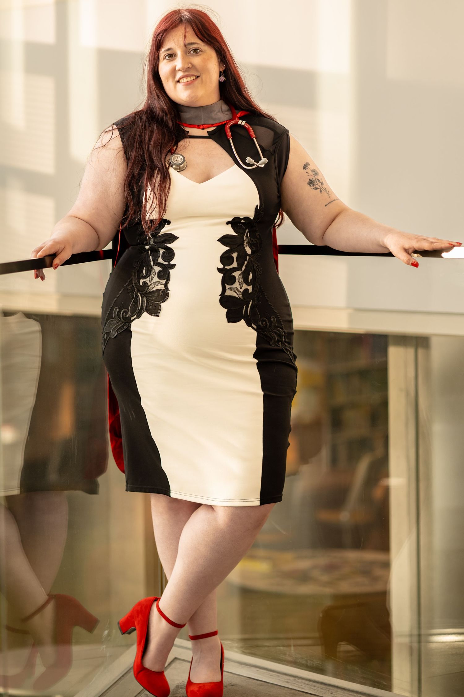 Woman in black and white dress with red heels and stethoscope posing confidently at a railing indoors.